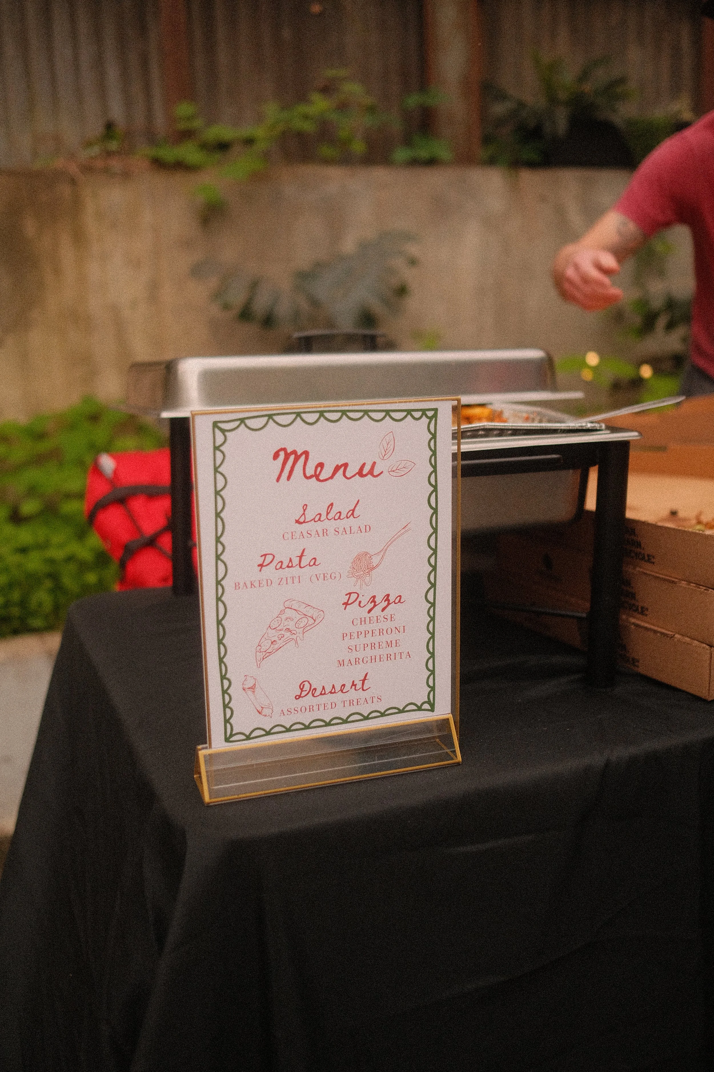 A menu stand on a black table listing various food options, including salad, pasta, pizza, and desserts. In the background, there is a person in a red shirt serving food from a stainless steel chafing dish.