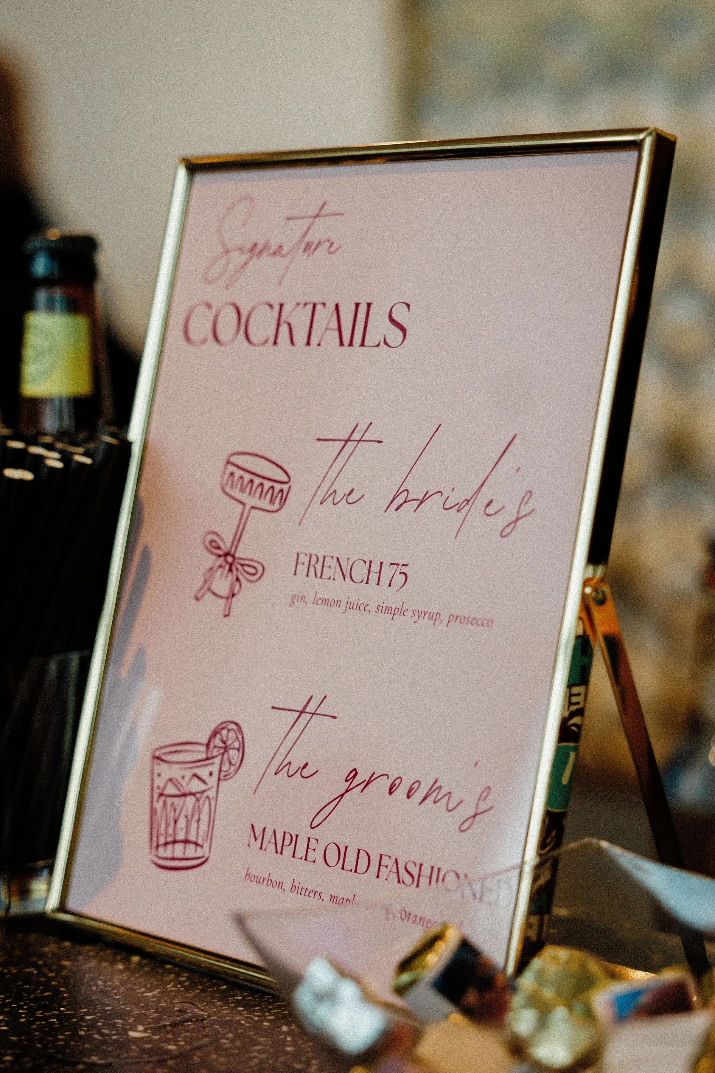 A photo of a sign listing signature cocktails, including French 75 with gin, lemon juice, simple syrup, and prosecco, and Maple Old Fashioned with bourbon, bitters, and maple syrup, with bottle of alcohol visible on the left side.