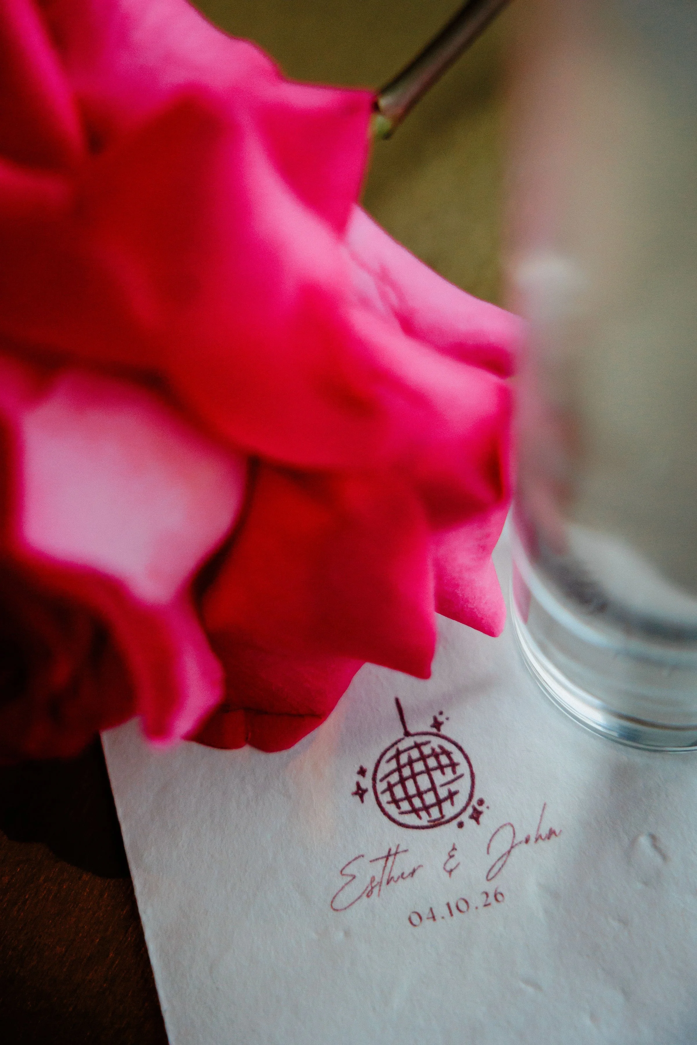 Close-up of a pink flower with a metallic stem, resting on a white piece of paper with red handwritten text, a holographic sticker, and a doodle of a disco ball.