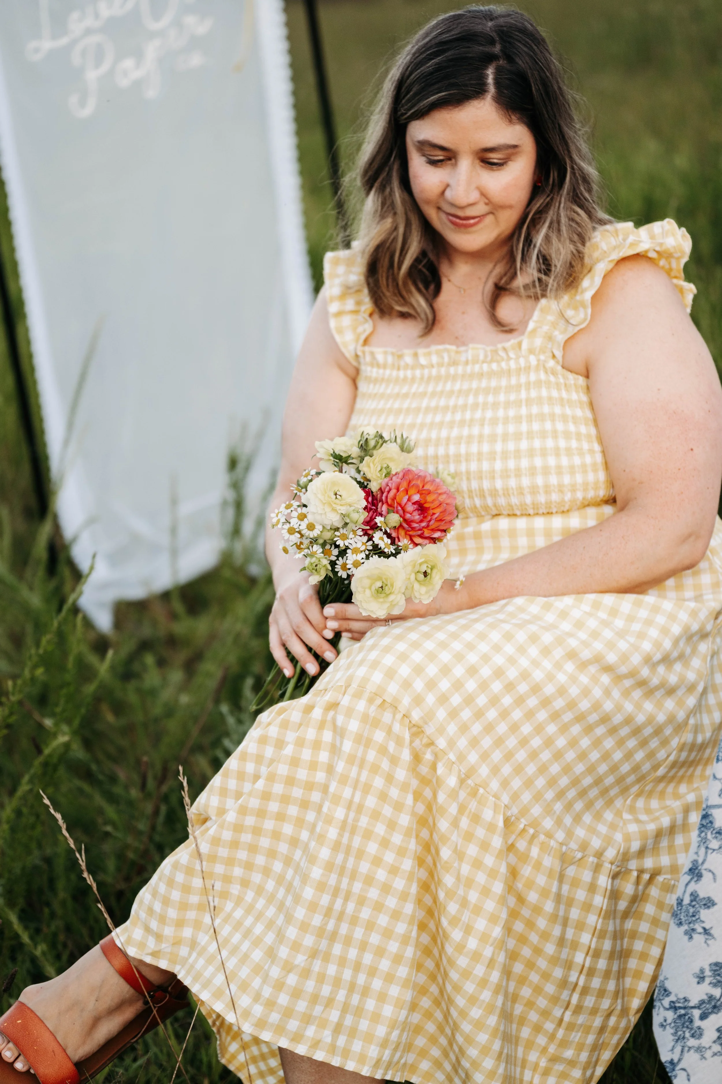 A woman in a yellow gingham dress sitting outdoors, holding a bouquet of flowers, with a grassy background and a white sign with partially visible writing behind her.
