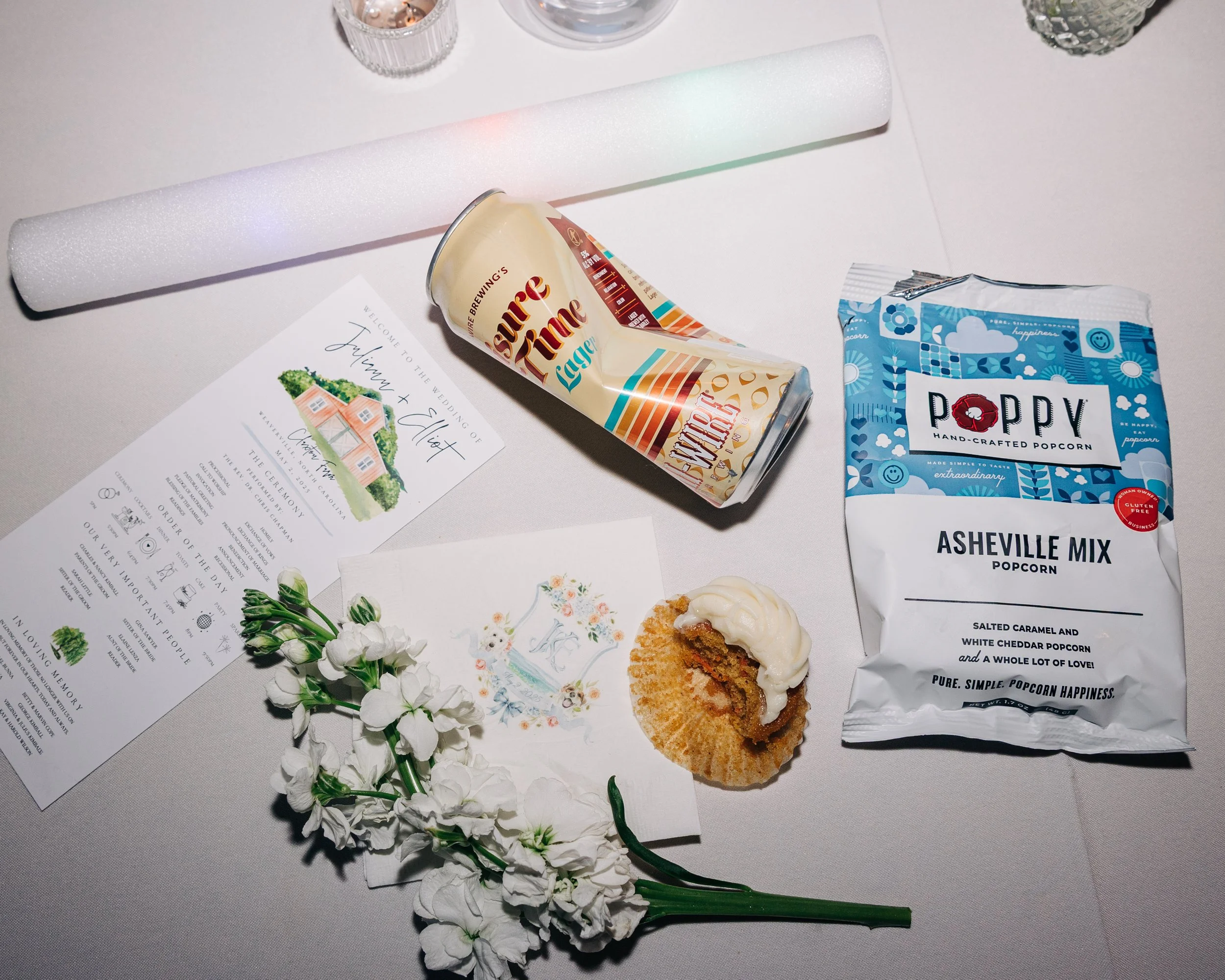 Wedding invitation, white flower sprig, can of Southern Time lager, bag of Poppy Asheville Mix popcorn, and a piece of dessert with white icing on a white tablecloth.