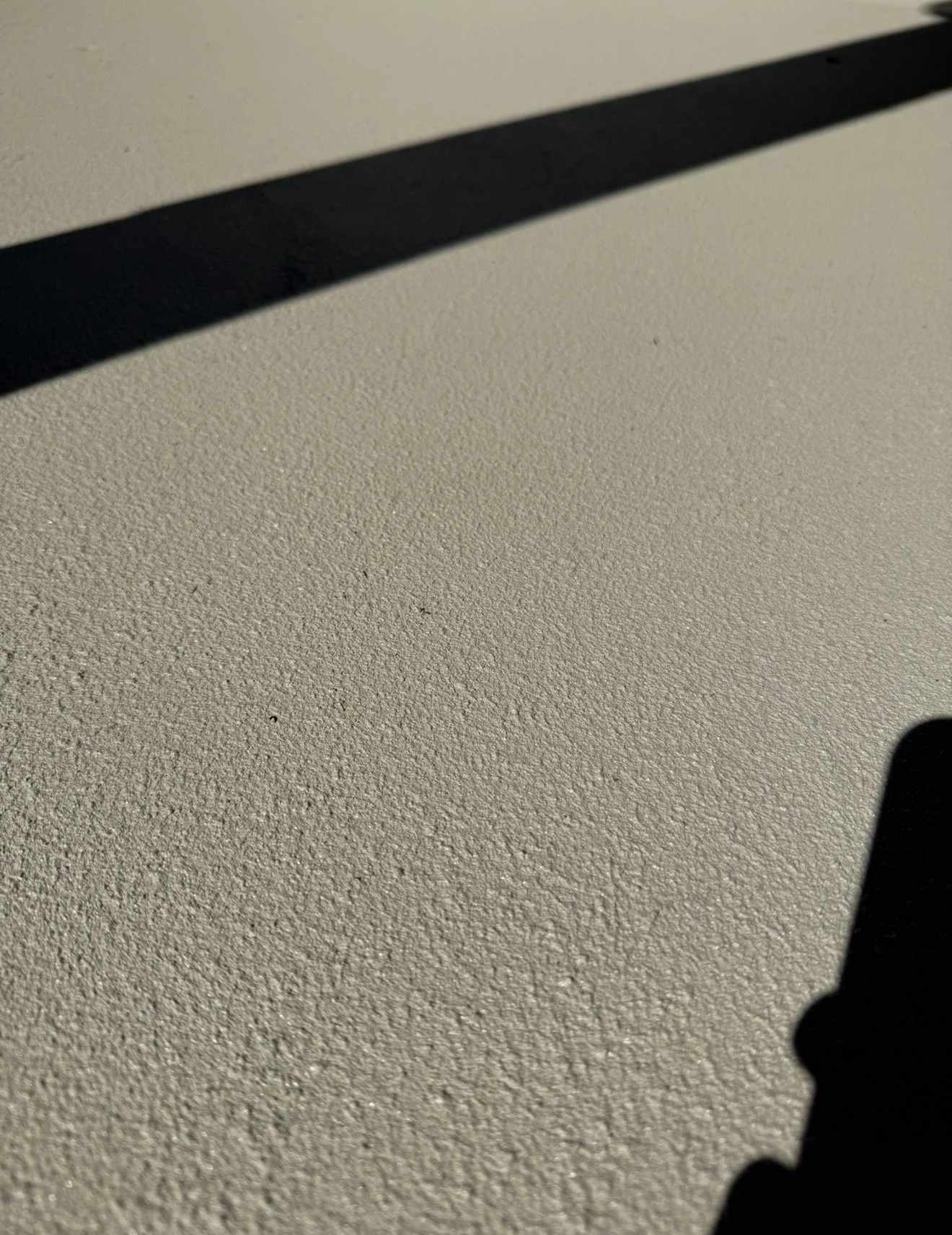 Close-up view of a shadow cast on a textured, beige epoxied floor with the edge of a dark object visible in the bottom right corner.