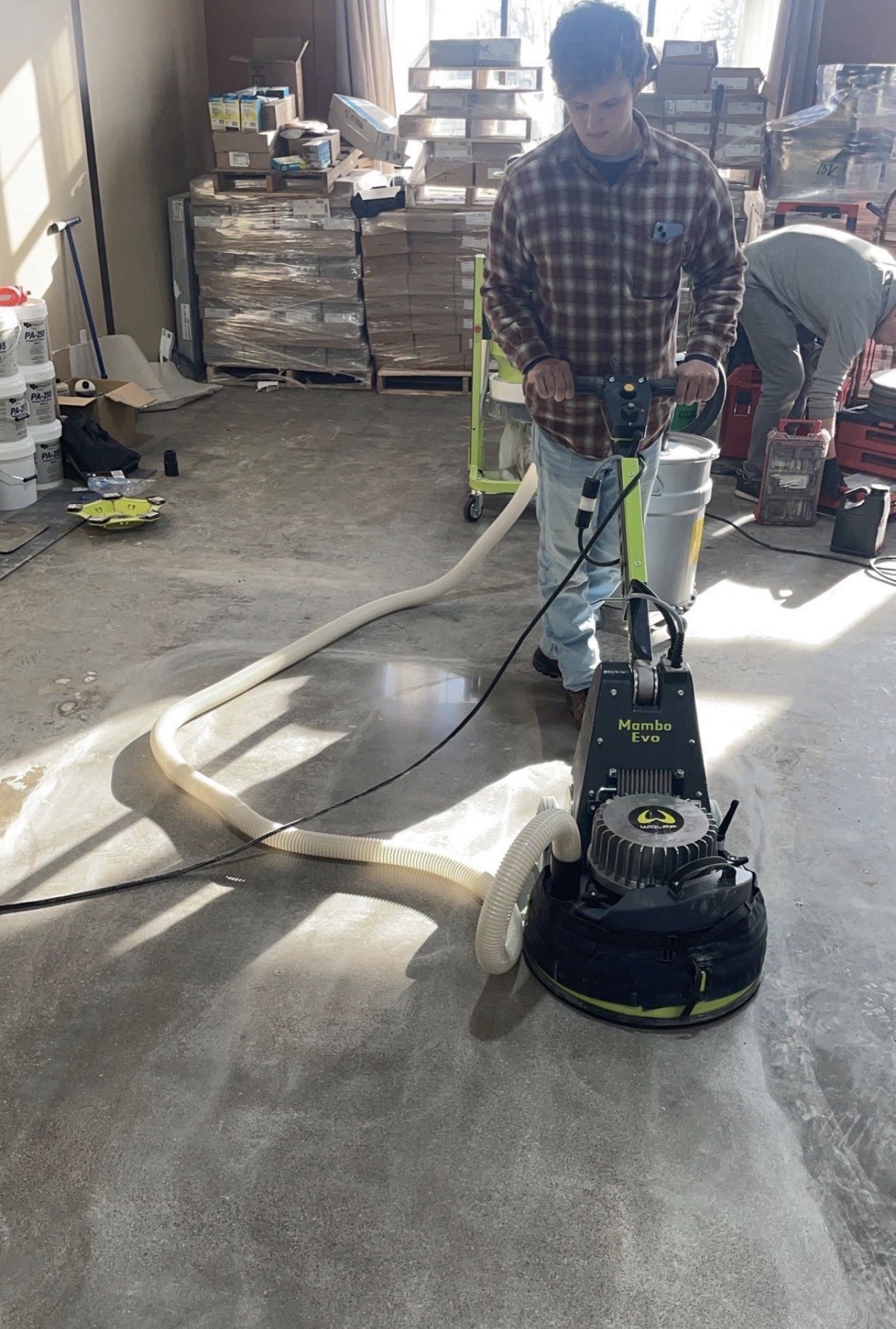 A man operating a floor grinding machine named Mambo Evo in a room. The room has stacks of boxes and supplies, with another person working in the background.