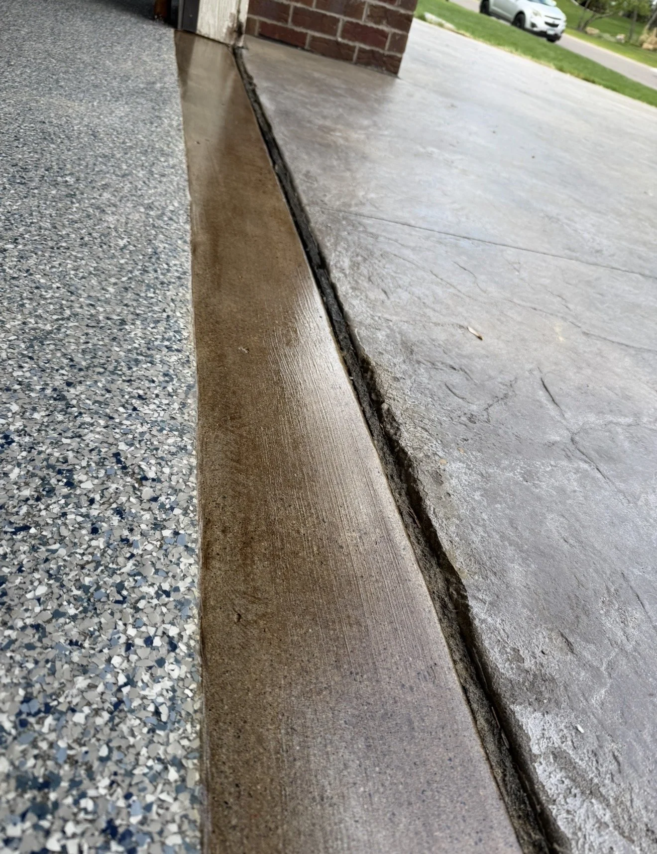 Close-up of a concrete sidewalk with a visible expansion joint, transitioning from a epoxy floor to a smooth stained and sealed surface, near the corner of a brick house with a grassy yard and a street with parked cars in the background.
