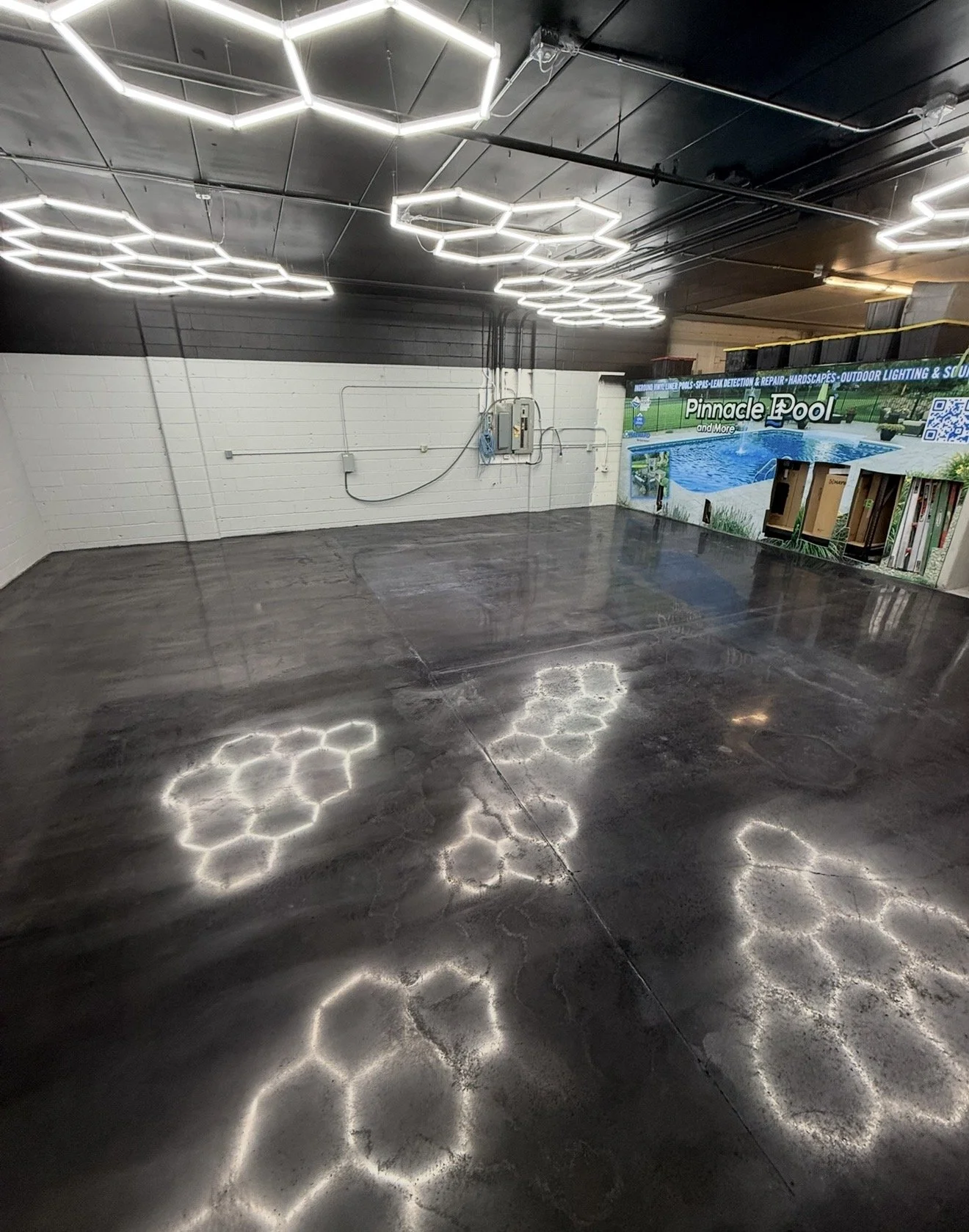 Empty indoor space with hexagonal lighting fixtures on the ceiling, a dark polished concrete floor reflecting the lights, and a wall with a Pinnacle Pool advertisement on the right side, featuring images of a swimming pool and outdoor landscape.