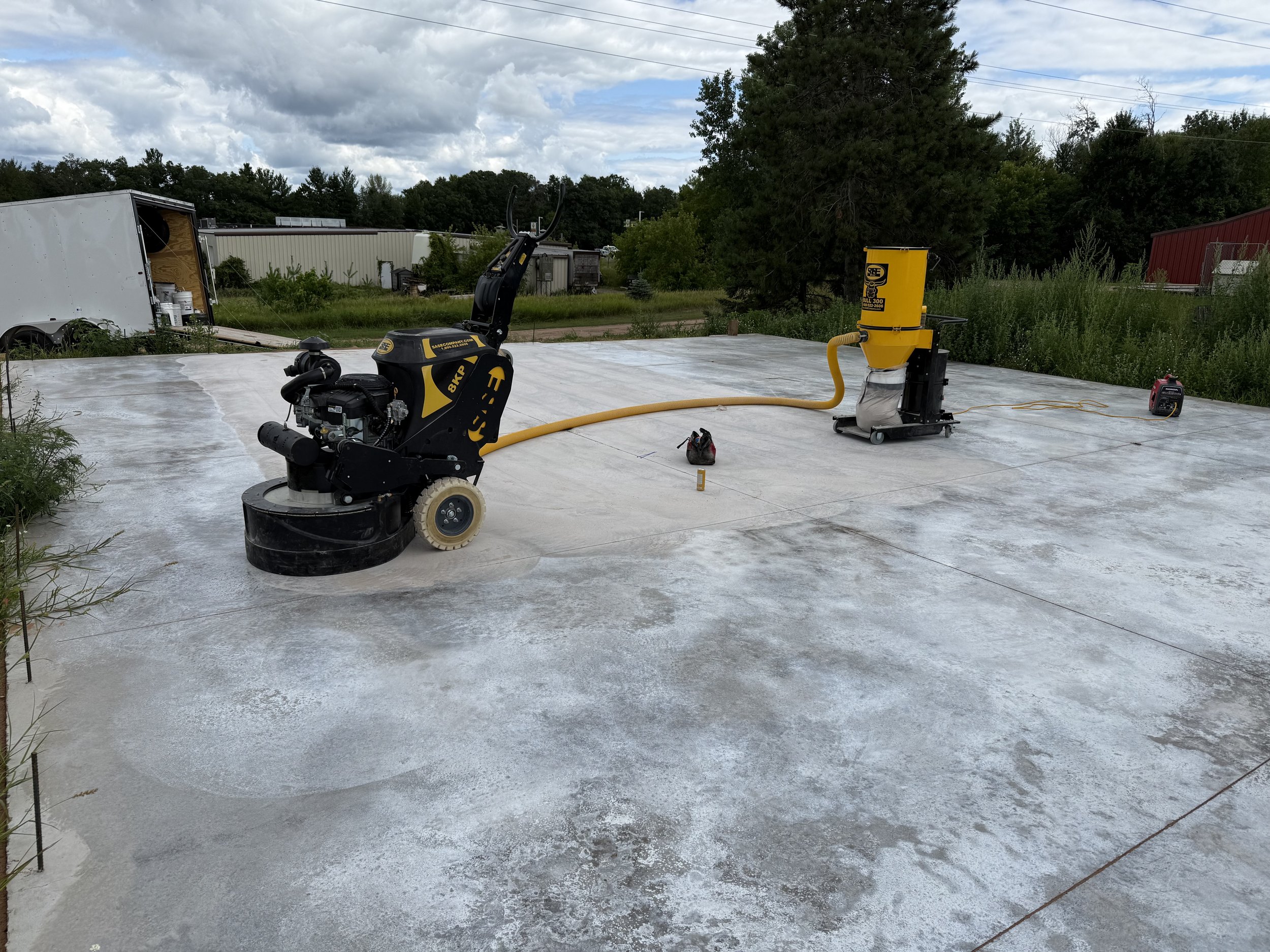 Construction site with a concrete slab, construction equipment including a floor grinder and a dust collector, and tools on the ground. Background features trees, a cloudy sky, and some buildings.