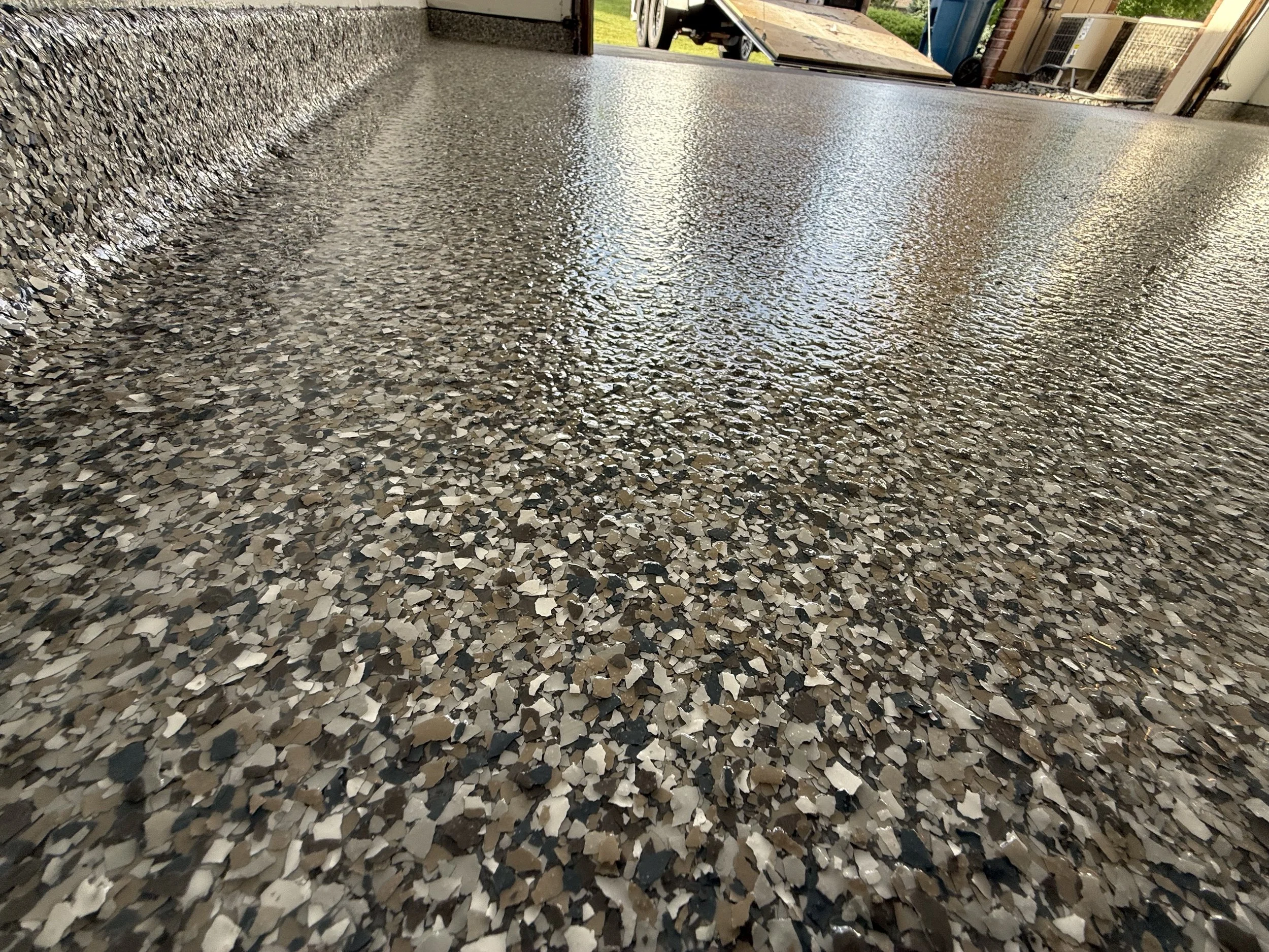 A freshly poured epoxy coated garage floor with a decorative mixed pebble and stone finish, located in a residential garage, with an open garage door leading outside.