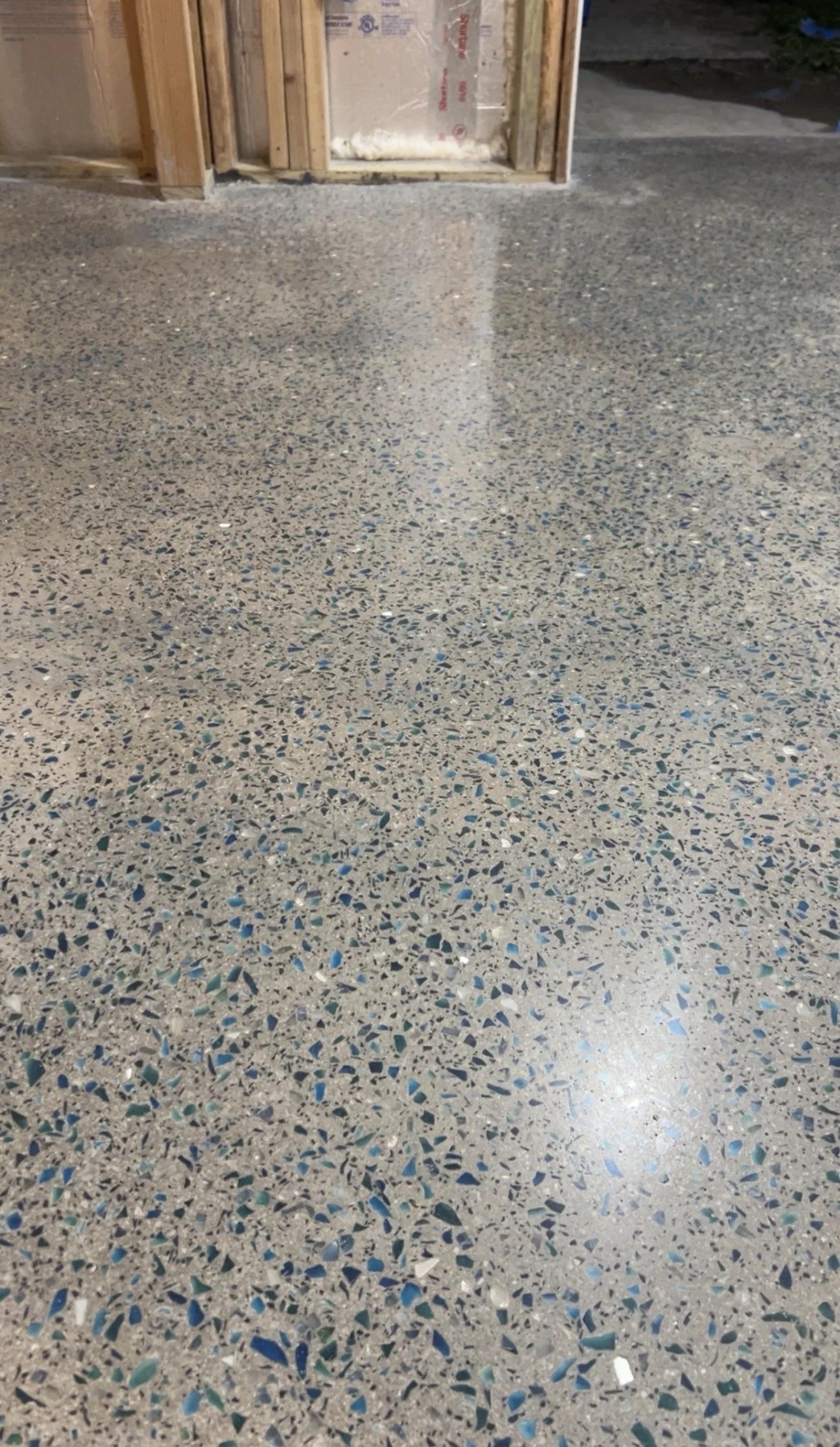 Newly poured epoxy floor with speckled pattern of blue, white, gray, and black chips, in a room under construction with exposed wooden framing and drywall. 