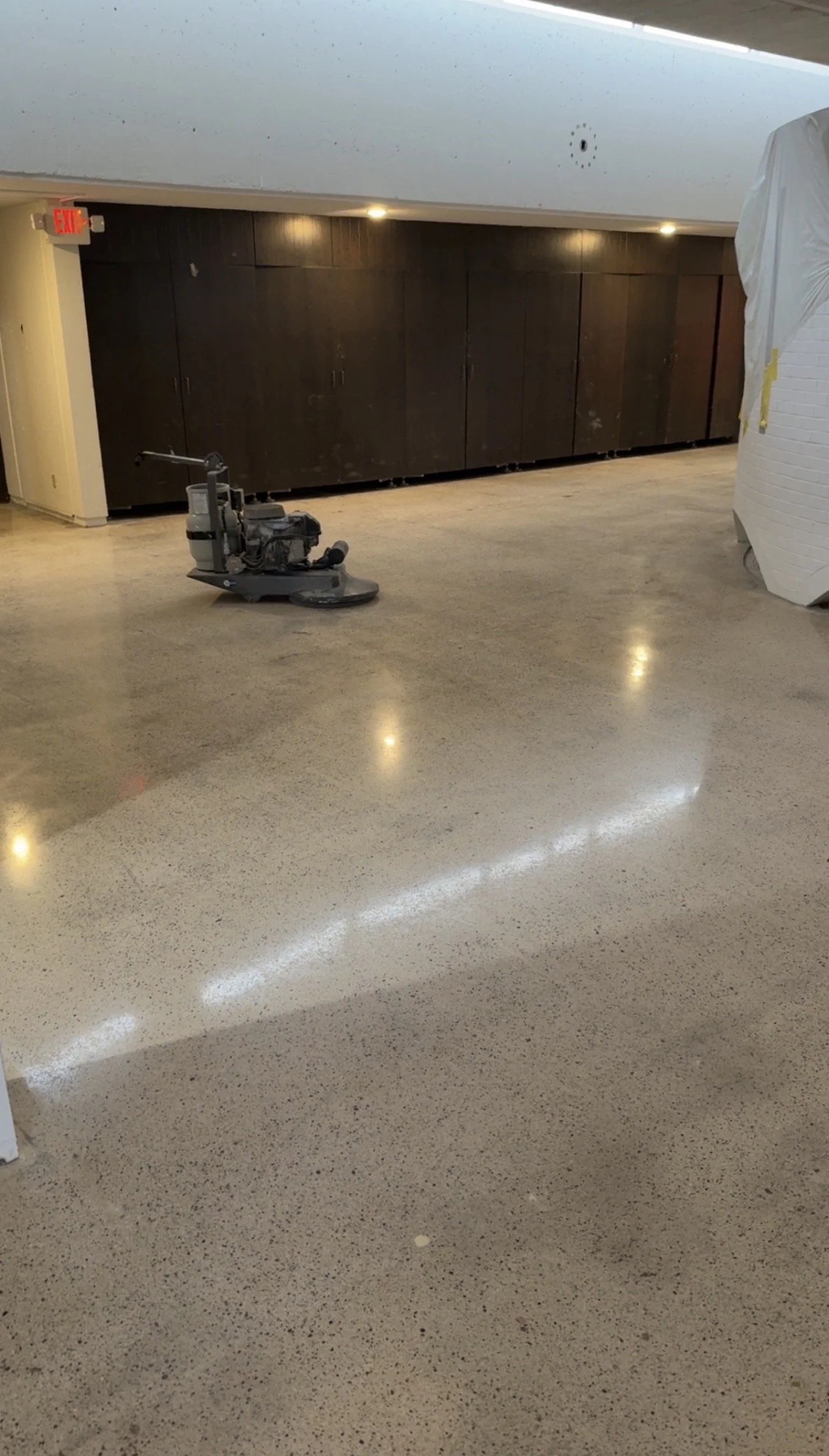 A floor scrubber machine on a polished concrete floor in an indoor space with dark wood cabinets, an exit sign, and a partially covered object.