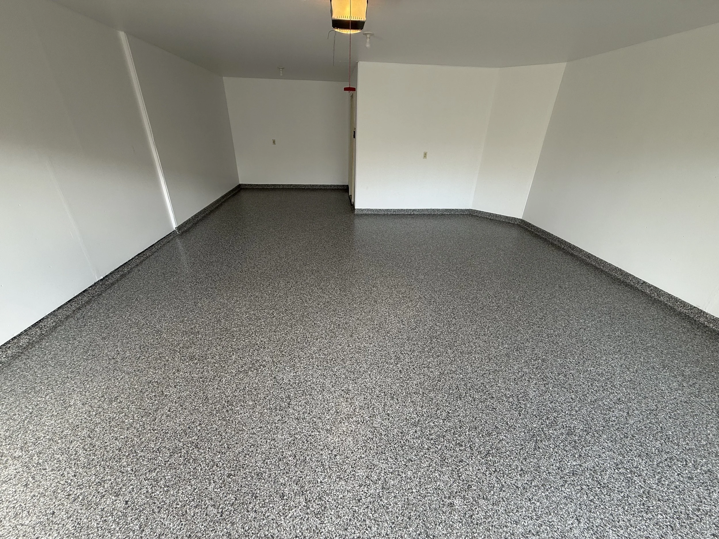 Empty room with white walls and gray speckled epoxy flake floor, ceiling light fixture, and a partially visible door.