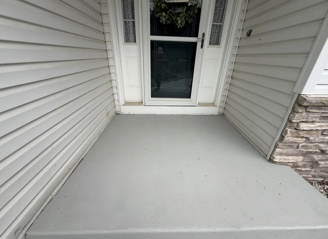 Empty concrete porch with an epoxied floor and white siding walls and a glass storm door with a wreath resting on the door.