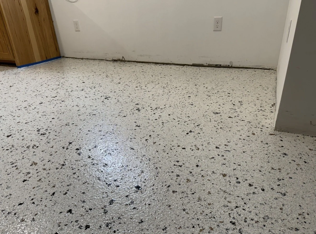 Image of a speckled white and black epoxy floor in a room with white walls, electrical outlets, and a section of wooden cabinetry.