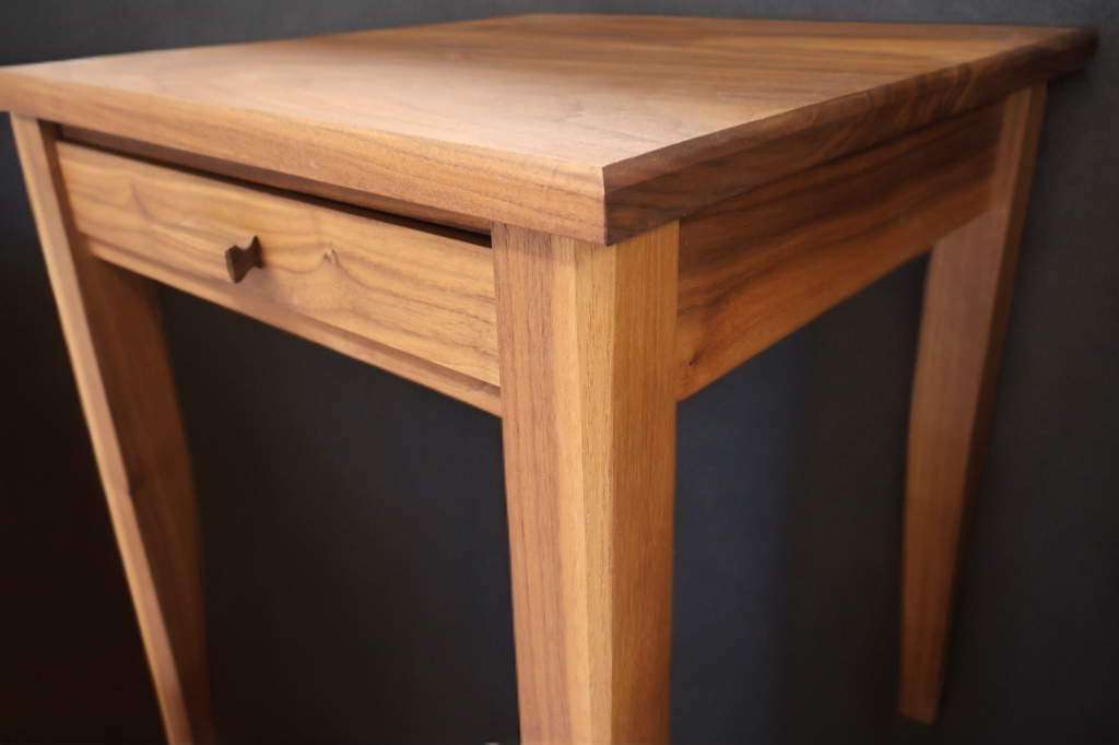 Wooden side table with a single drawer, set against a black background.