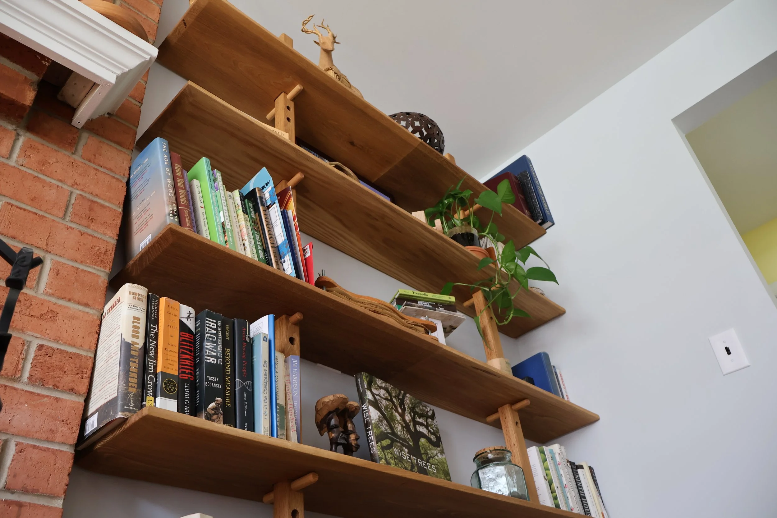 Wooden bookshelves mounted on a wall with various books, plants, decorative items, and a small sculpture on the shelves, with a brick wall and ceiling visible.