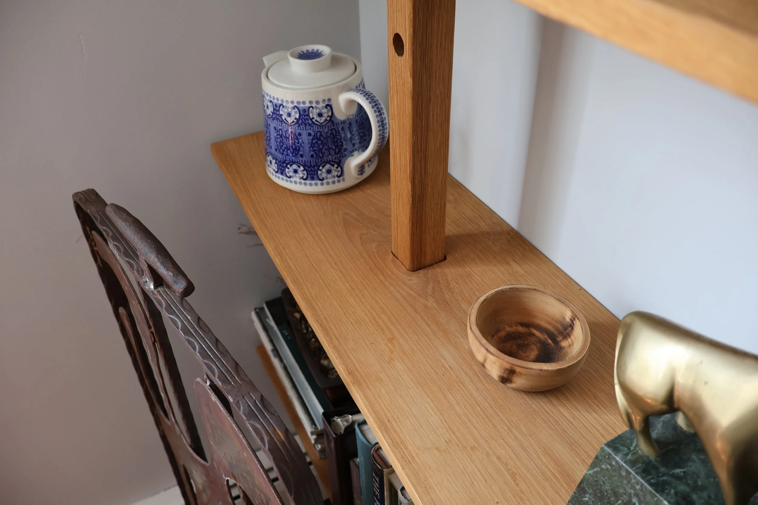 Wooden shelving unit with a decorated teapot, a wooden bowl, and a ceramic item with gold accents. Part of a stove and a wooden chair are visible in the background.