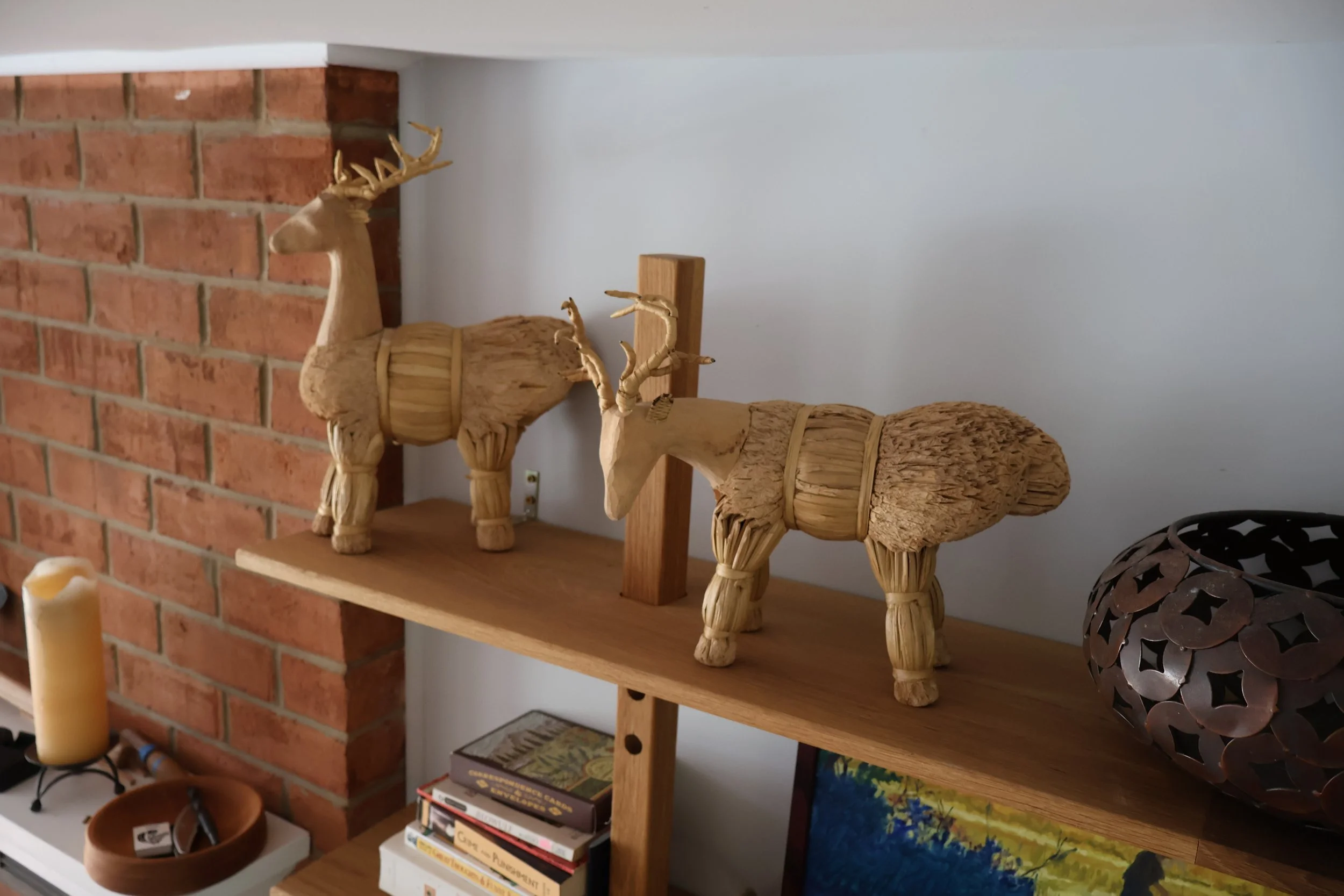 Wooden deer figurines on a wooden shelf next to a brick fireplace and a decorative lantern, with candles and books below.