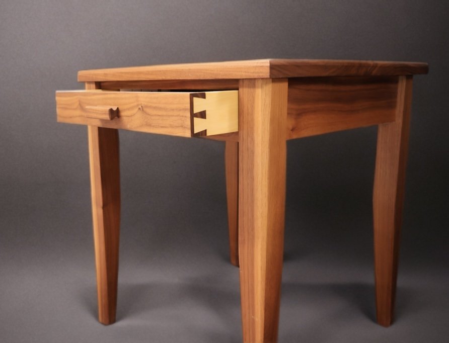Wooden side table with a single drawer and tapered legs against a dark background.