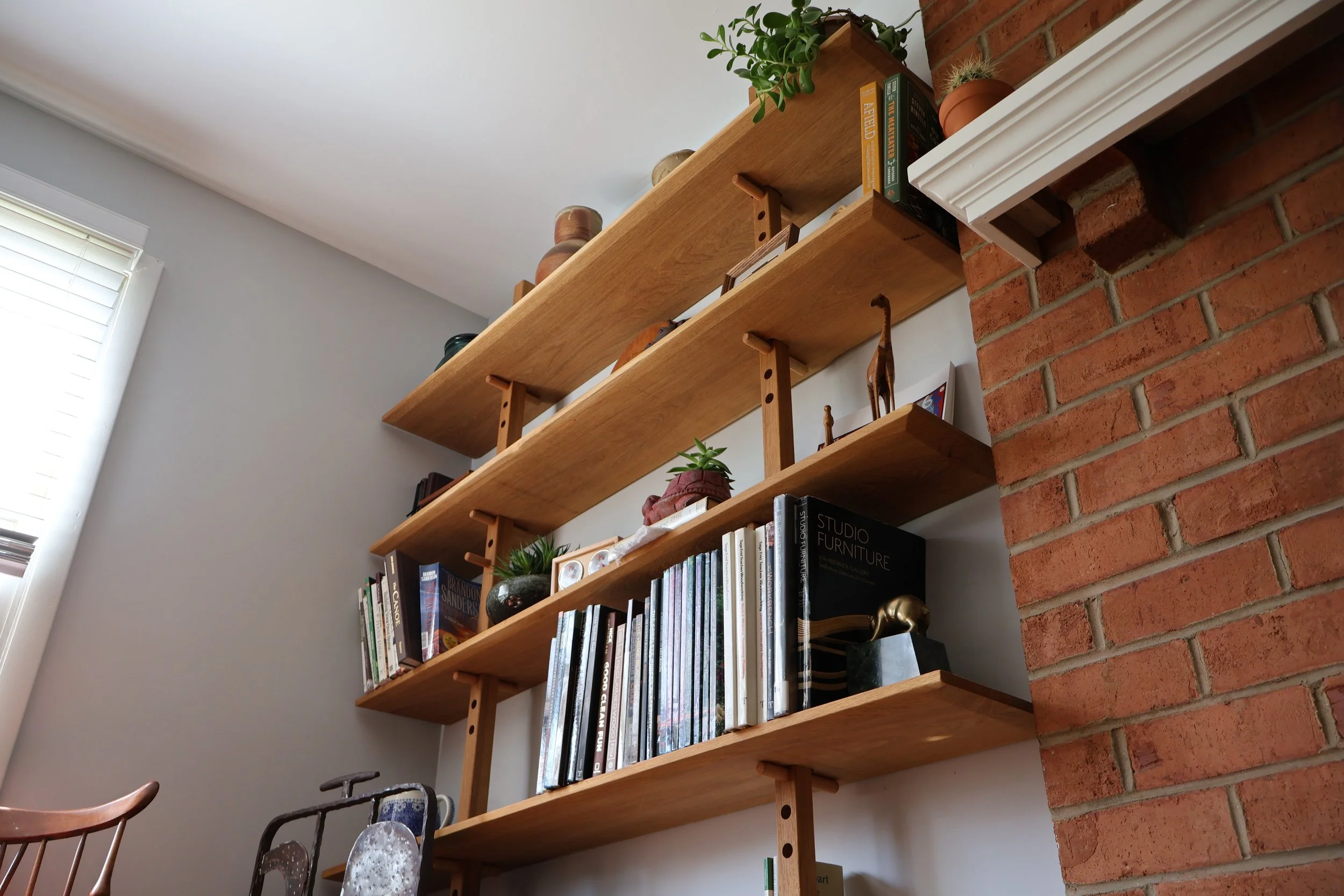 A wooden bookshelf mounted on a wall with books, small potted plants, and decorative items, next to a brick wall in a cozy living space.