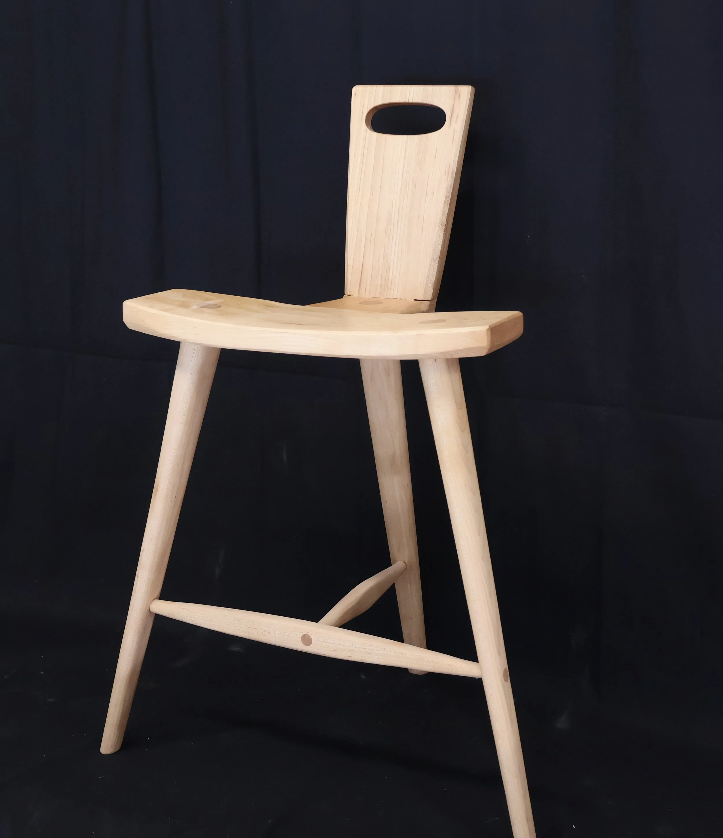 A light-colored wooden chair with a curved seat, four angled legs, and a tall backrest with an oval cutout, set against a black background.
