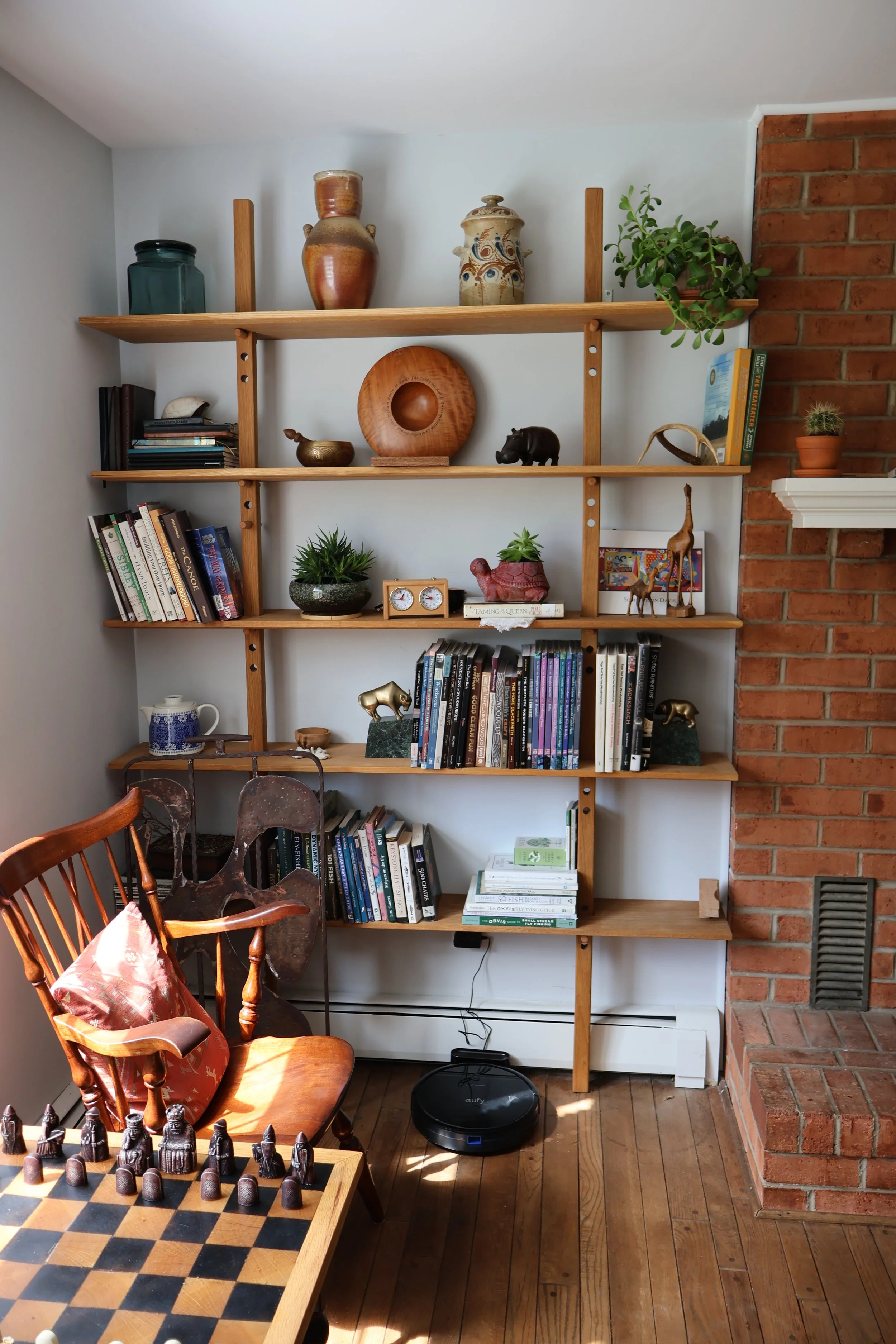 Interior view of a room featuring a wooden bookshelf filled with books, decorative objects, and plants. There is a table with a chessboard and chess pieces in the foreground. An armchair with a pink patterned pillow, a robot vacuum cleaner on the flo