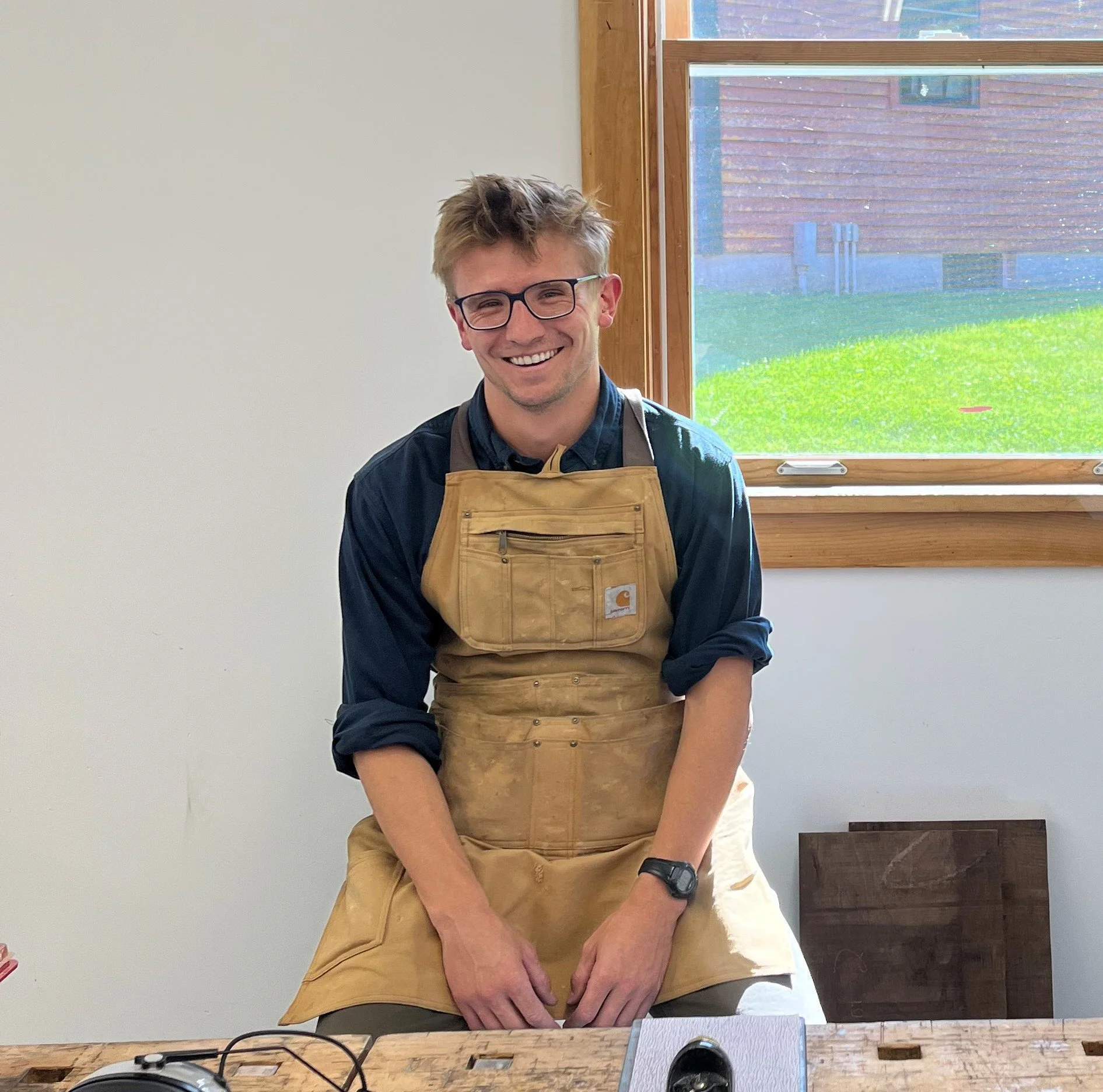 A young man with glasses, messy light brown hair, wearing a dark long sleeve shirt and a tan apron, smiling while standing in a room with a large window showing a green yard outside.