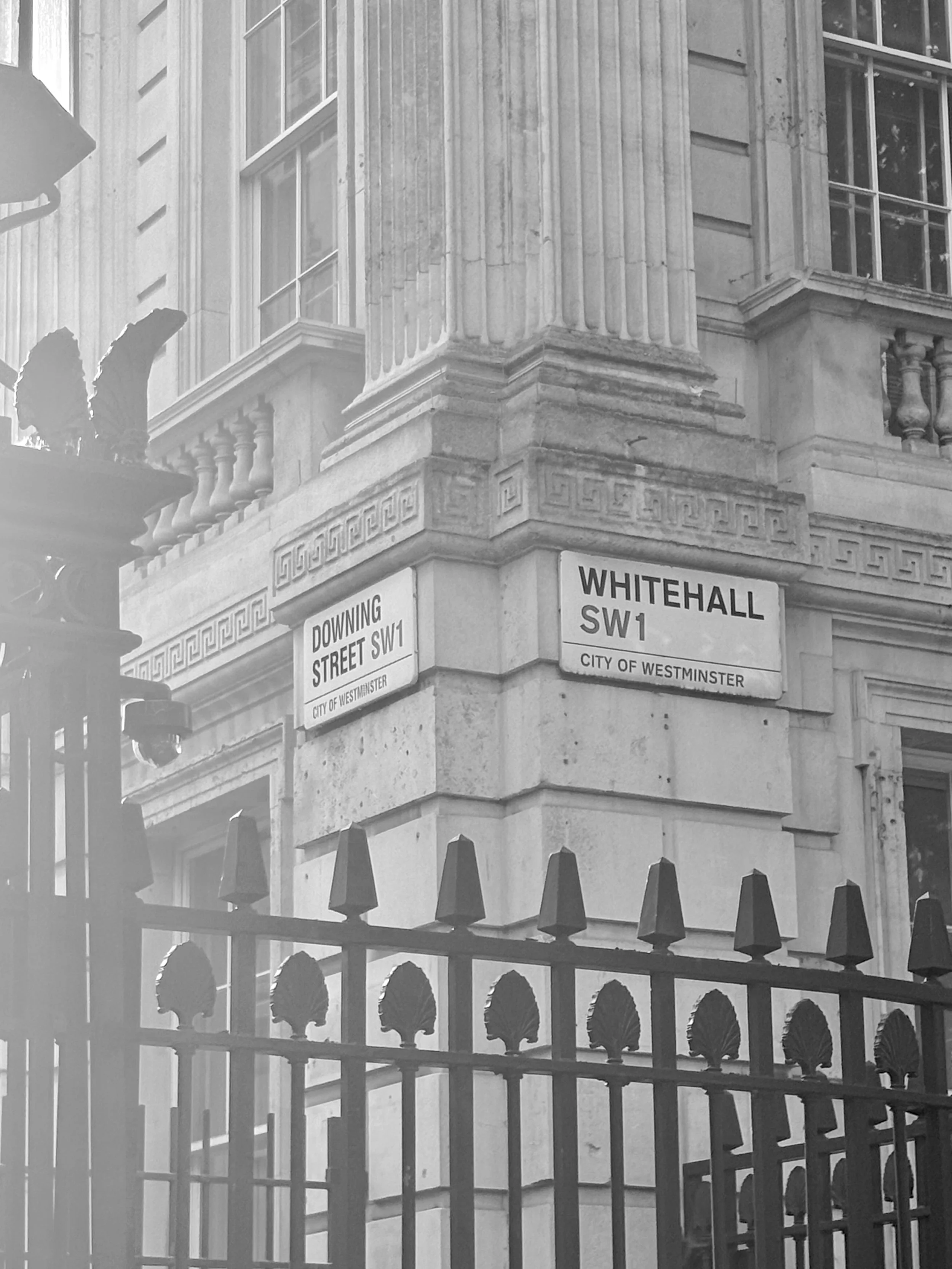 Street corner of a historic building with classical architecture, ornamental cornice details, and street signs for Downing Street and Whitehall in Westminster, London, seen behind a decorative iron fence.