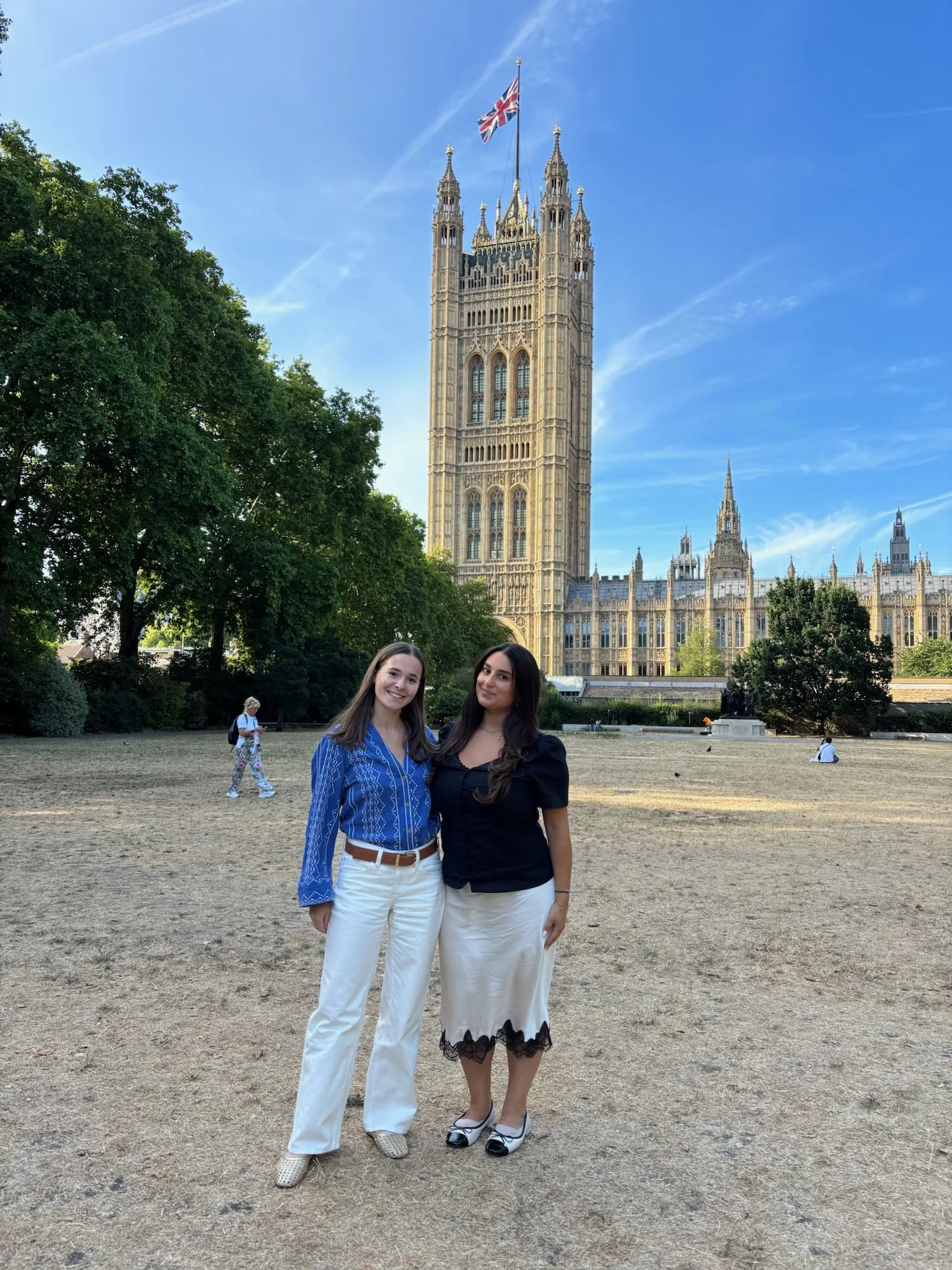 Two young women standing in front of the UK Parliament building in London, smiling at the camera. One is wearing a blue patterned blouse and white pants, and the other is wearing a black top and white skirt with black lace trim. There are trees and a few people in the background.