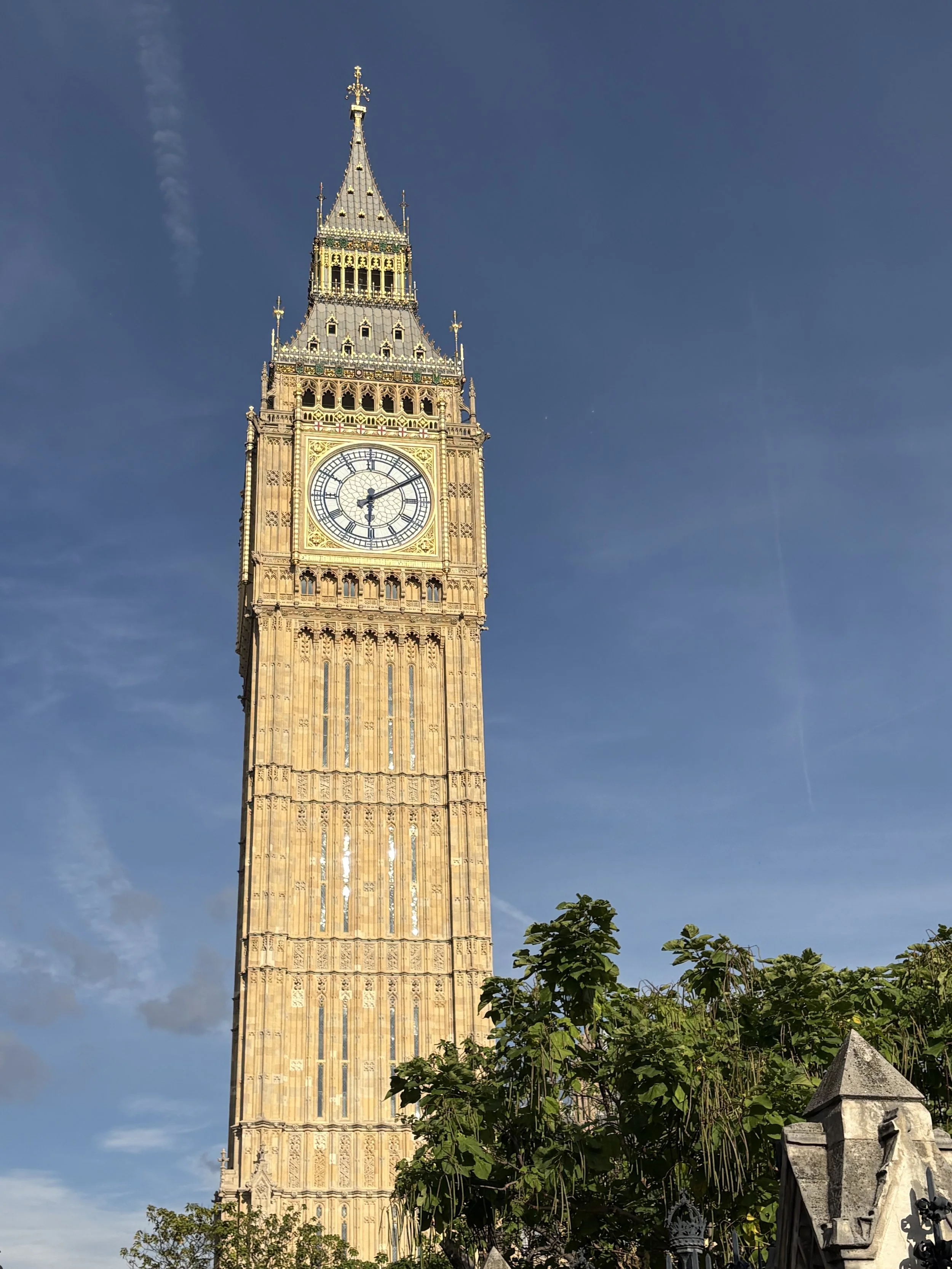 The clock tower known as Big Ben in London, England, with a clear blue sky in the background and greenery in the foreground.