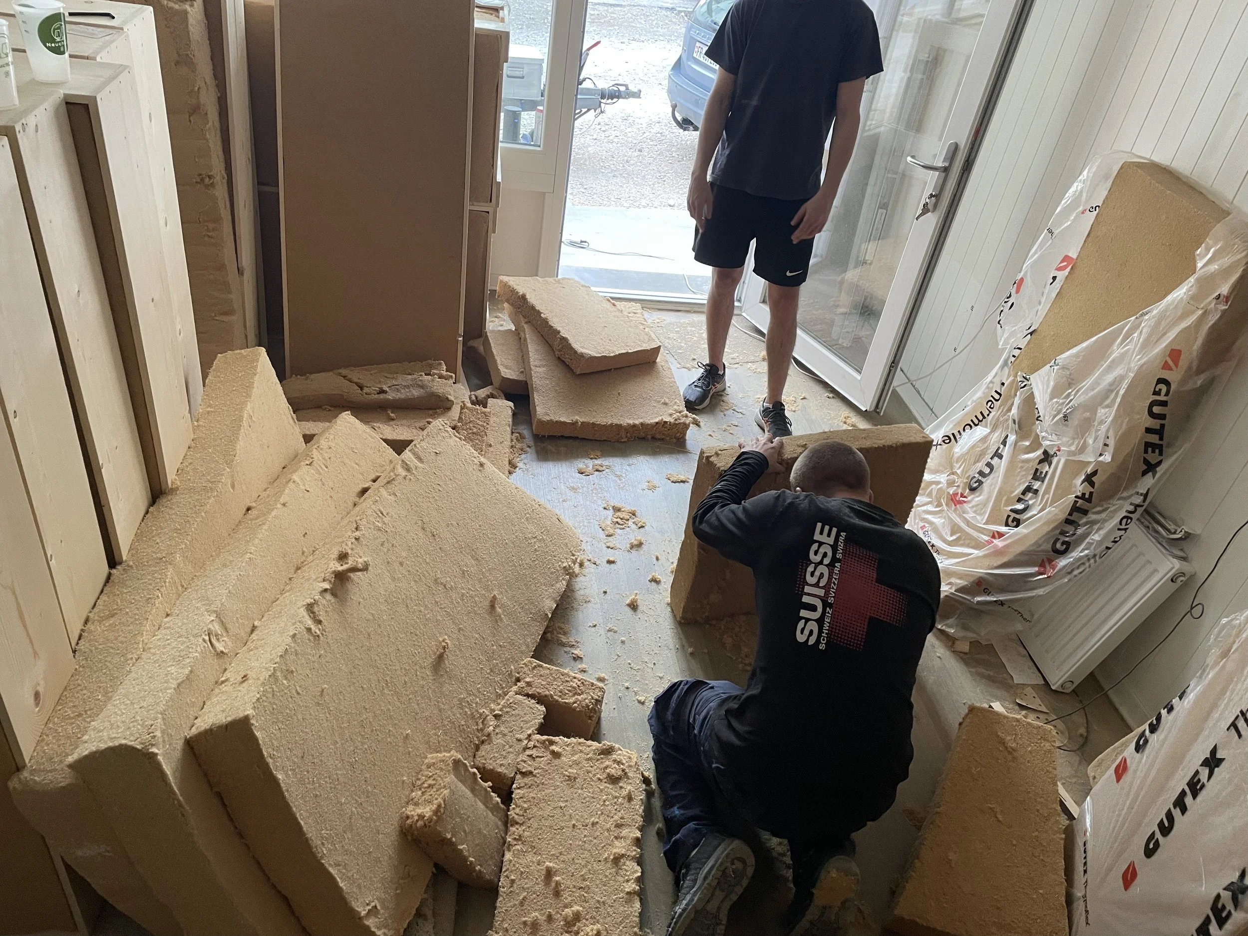 Deux personnes travaillent à l'intérieur d'une pièce en construction ou en rénovation, entourées de blocs d'isolation en polystyrène. Une personne est debout, portant un t-shirt noir et un short noir, tandis qu'une autre est accroupie, manipulant un 