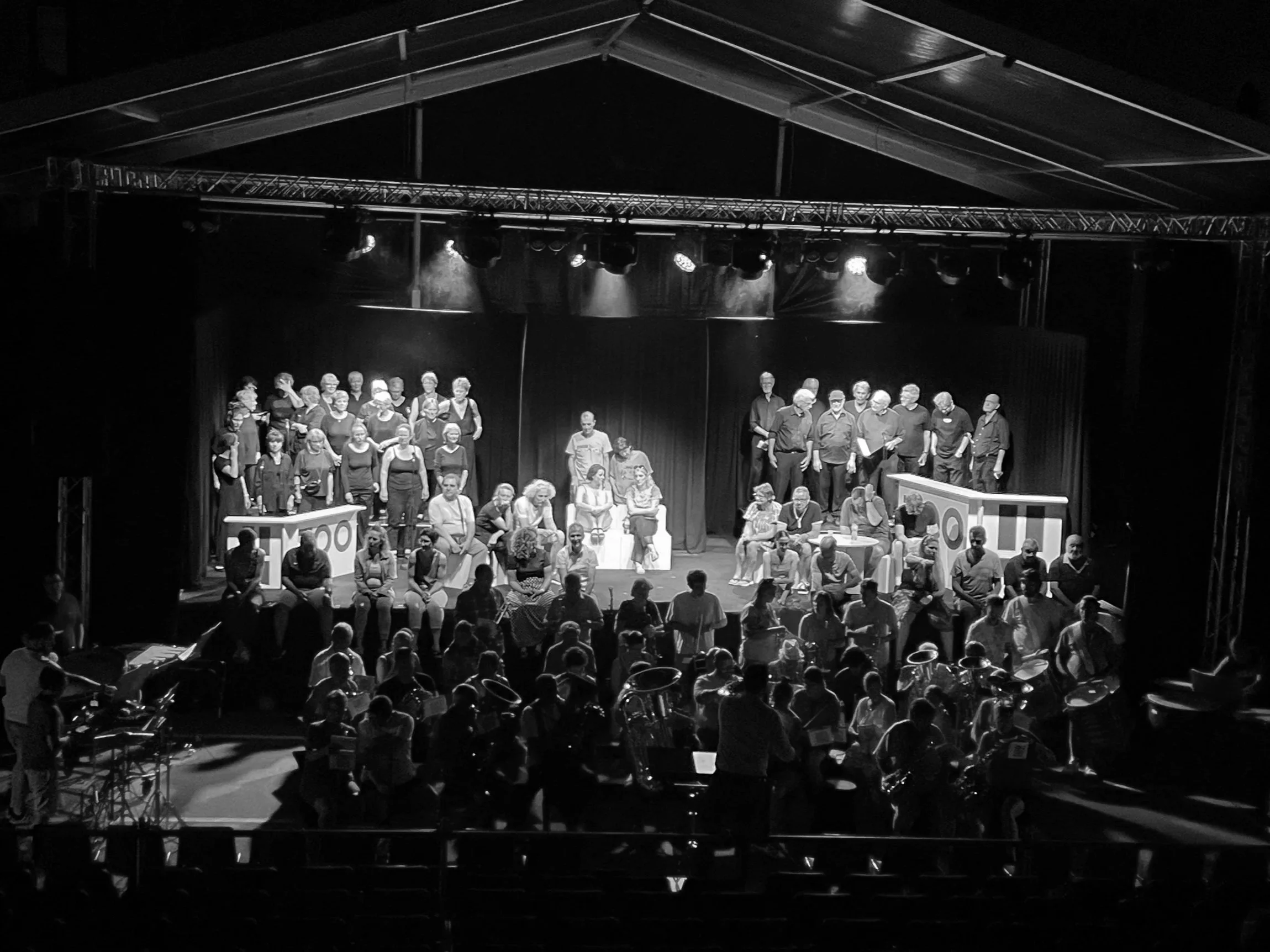 Une scène de théâtre avec plusieurs personnes sur scène et dans la salle, éclairage de scène, public assis et musiciens en avant-plan.