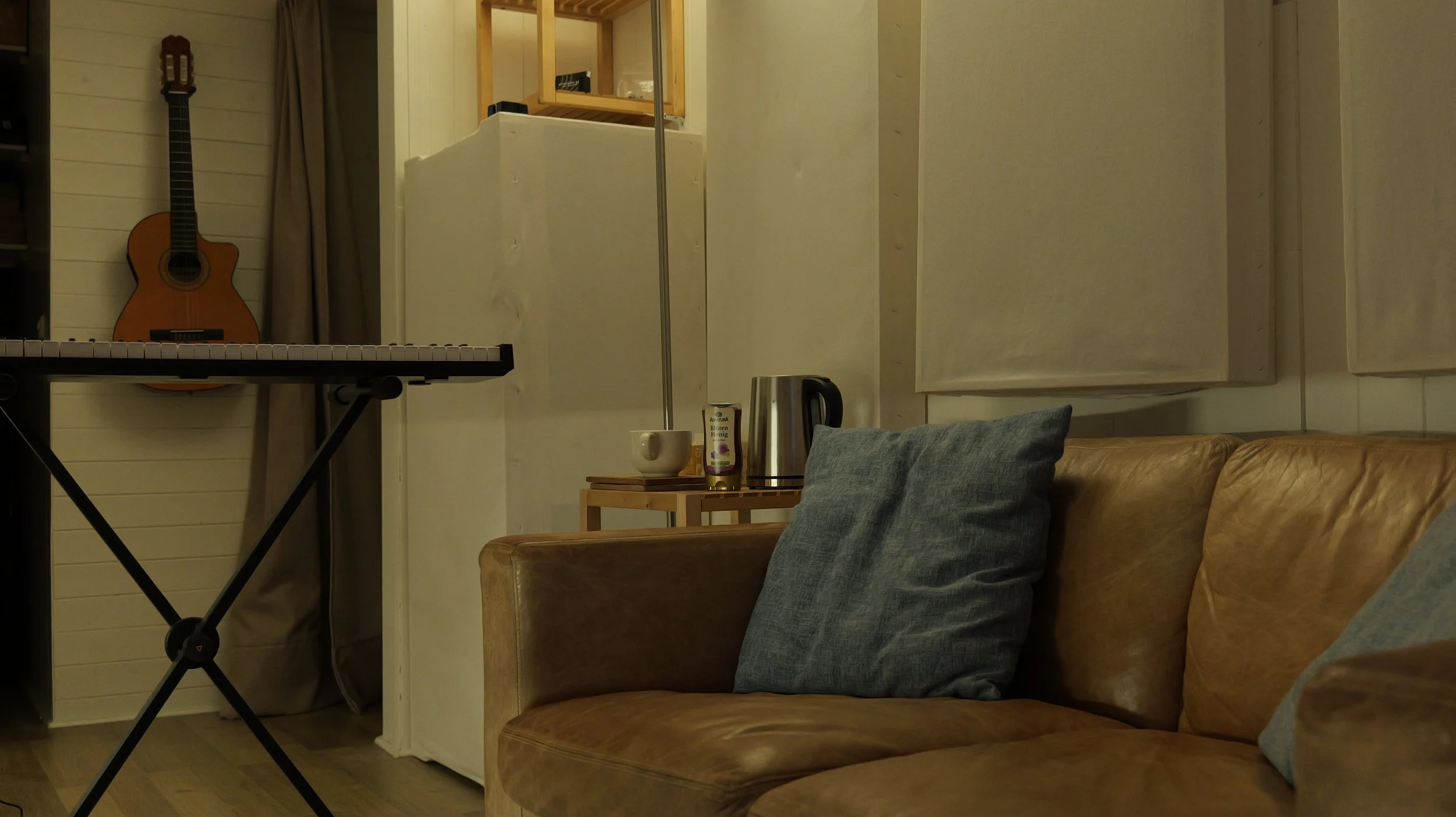 Un salon avec un canapé en cuir beige, des coussins gris, une guitare sur un mur en bois clair, une petite table avec une tasse, une bougie, une bouilloire électrique et une boîte de thé ou tisane.