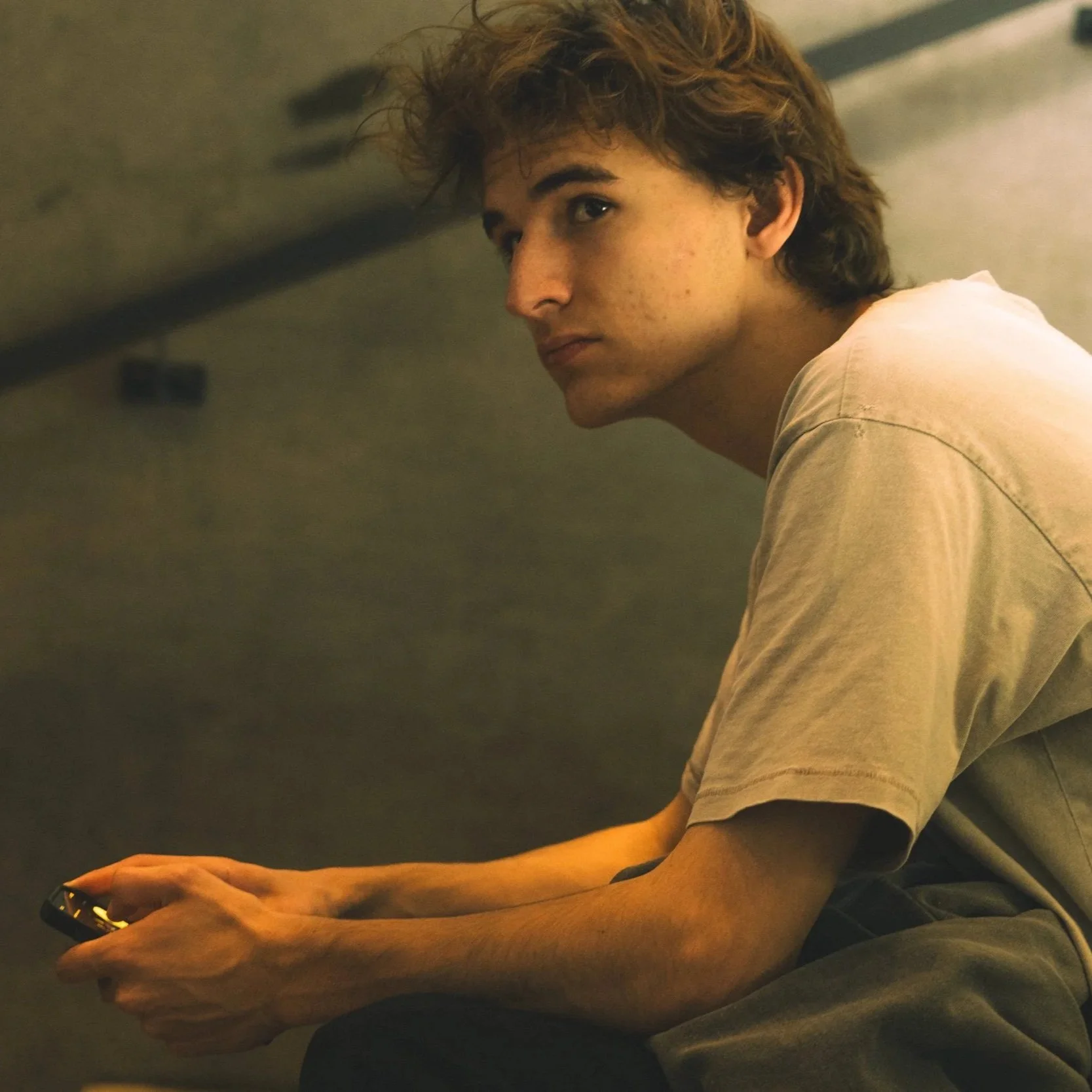 Un jeune homme aux cheveux bouclés, portant un t-shirt beige, est assis dans un endroit sombre, regardant par-dessus son épaule et tenant un téléphone portable dans sa main gauche.