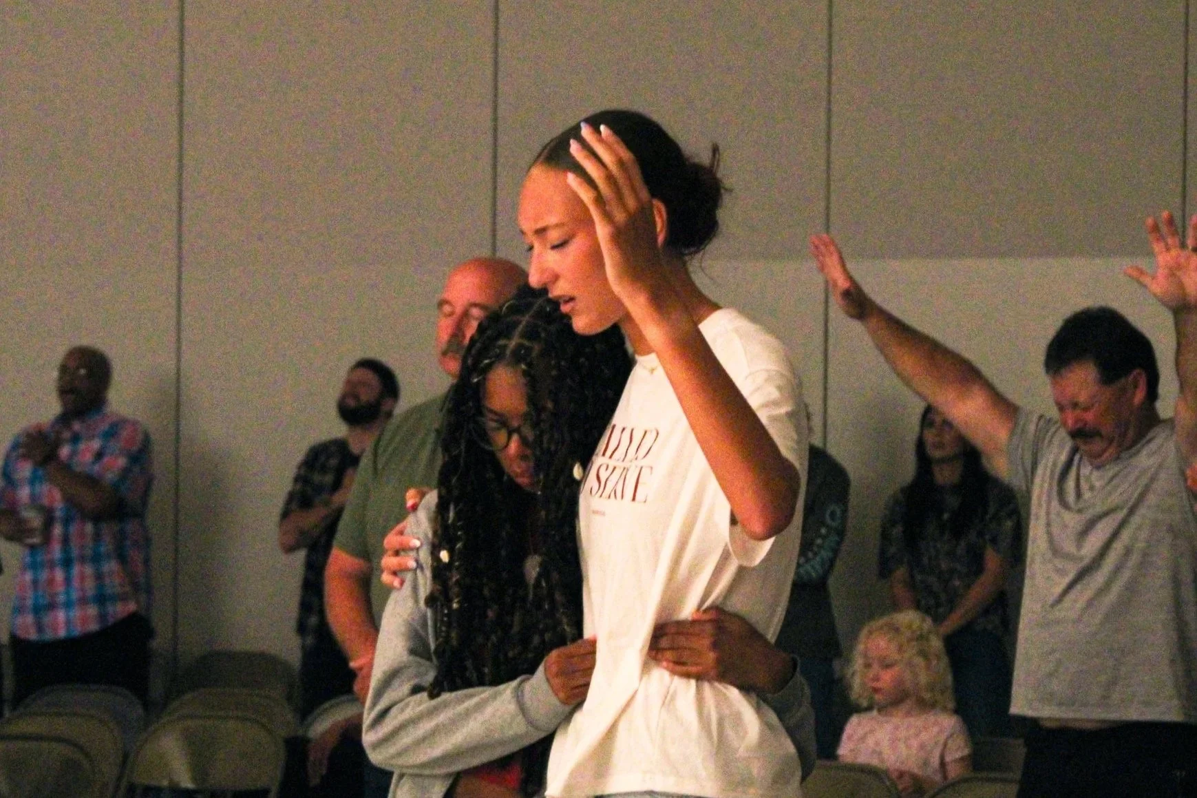 People embracing and praying in a church or conference room, with some individuals raising hands and others with eyes closed in prayer.