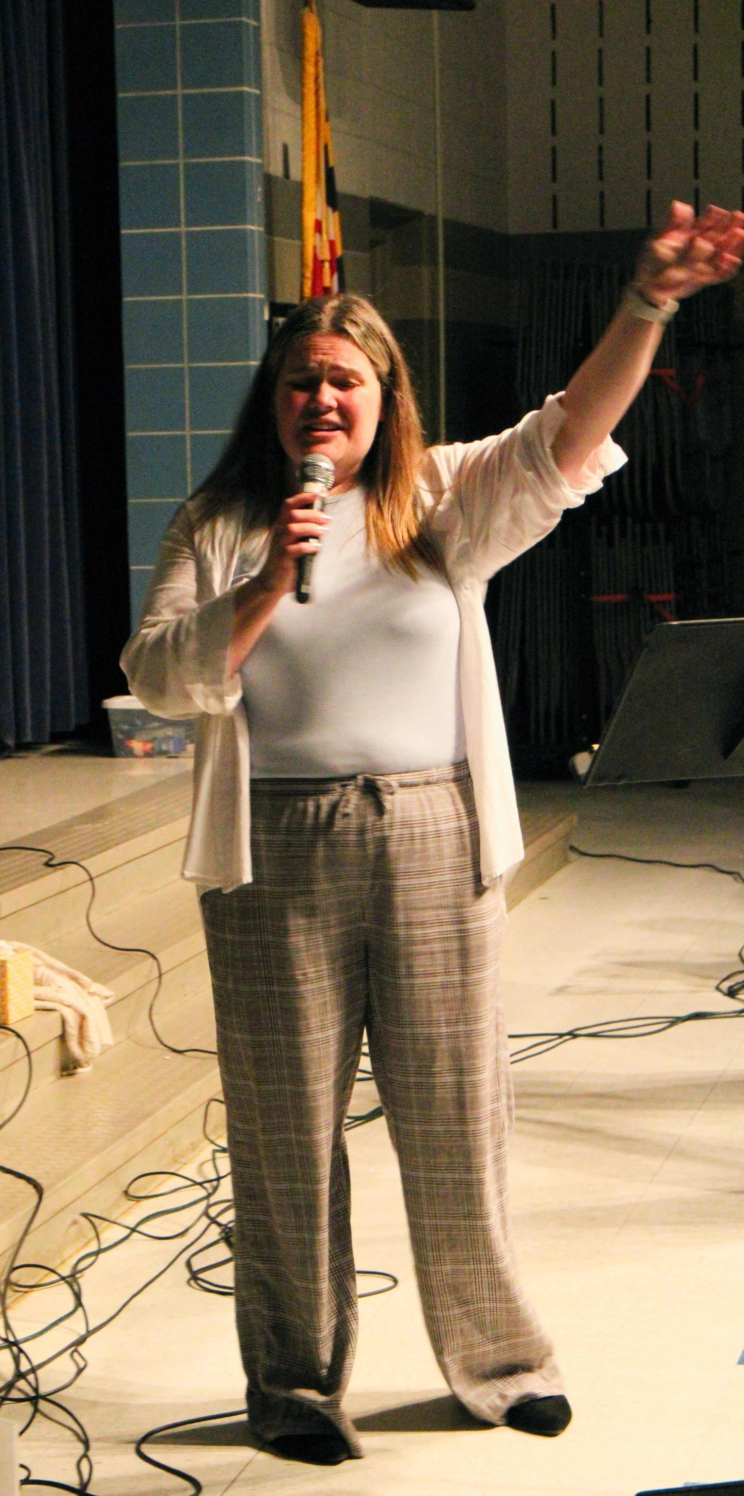 A woman with long hair holding a microphone, gesturing with her right arm, speaking on stage in a room with blue tiled walls and a flag in the background, wearing plaid pants and a white top.