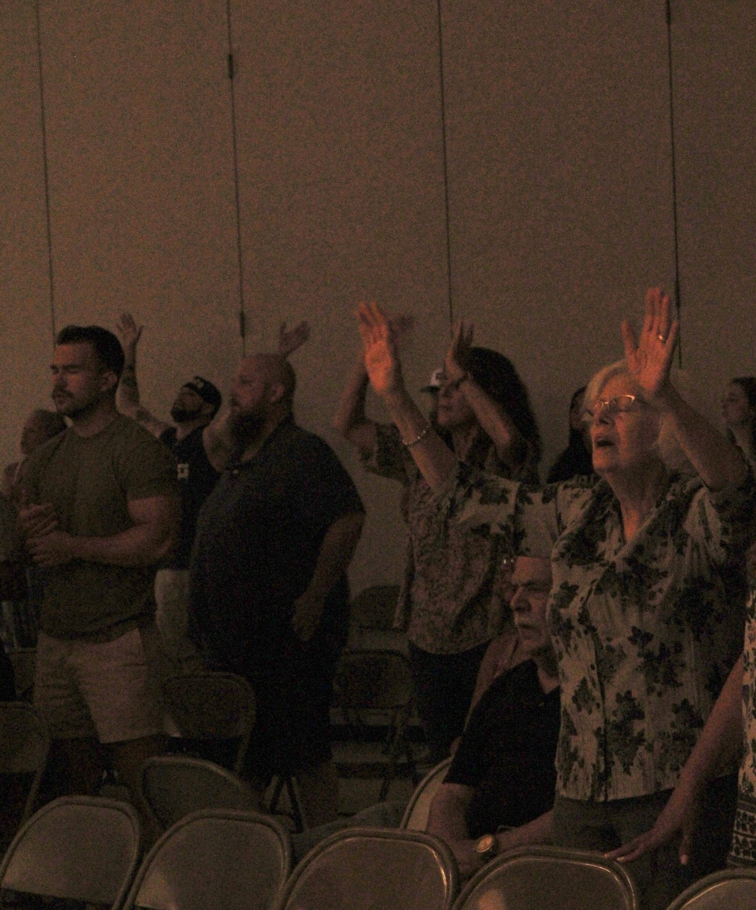 People seated in a dark room, raising their hands, possibly during a presentation or event. The wall behind them is plain and dark.