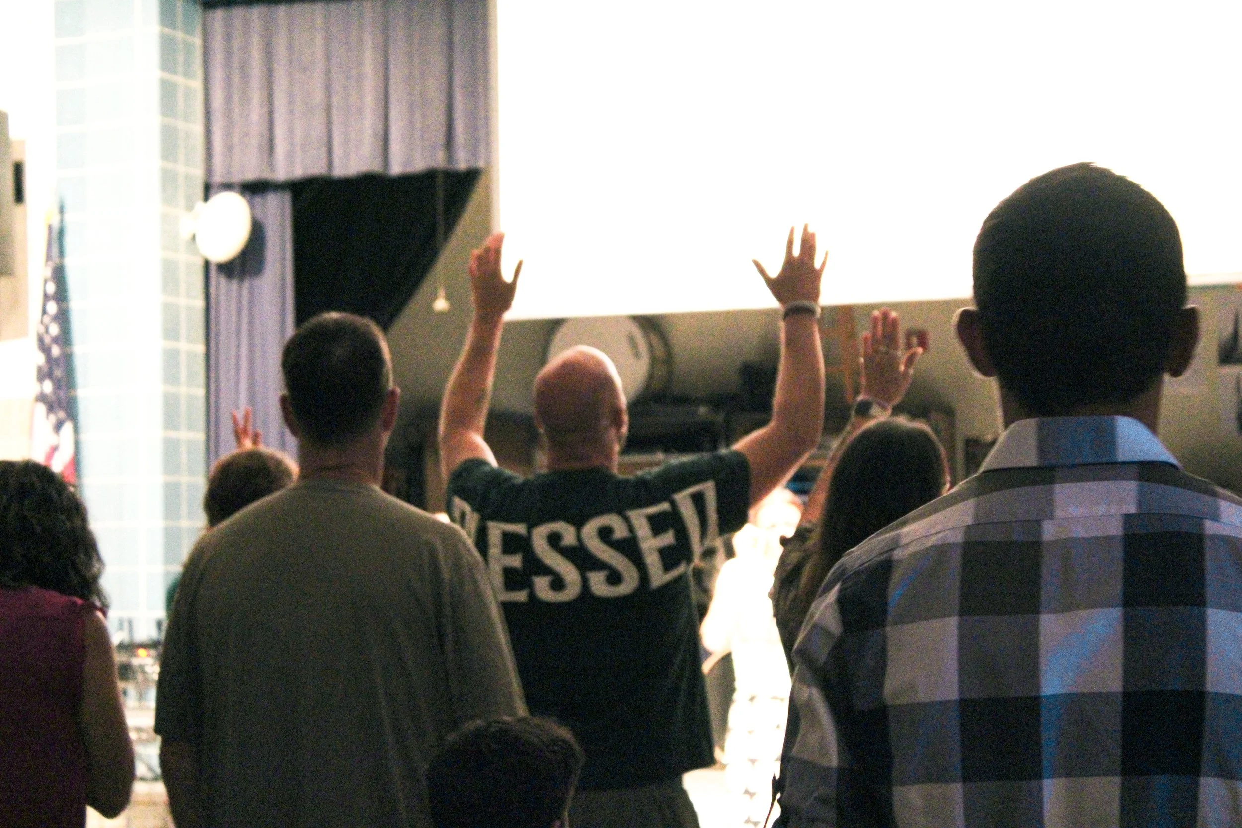 People attending a conference or presentation, with some raising their hands in response to a speaker or question on stage, in a large room with a bright projection screen.