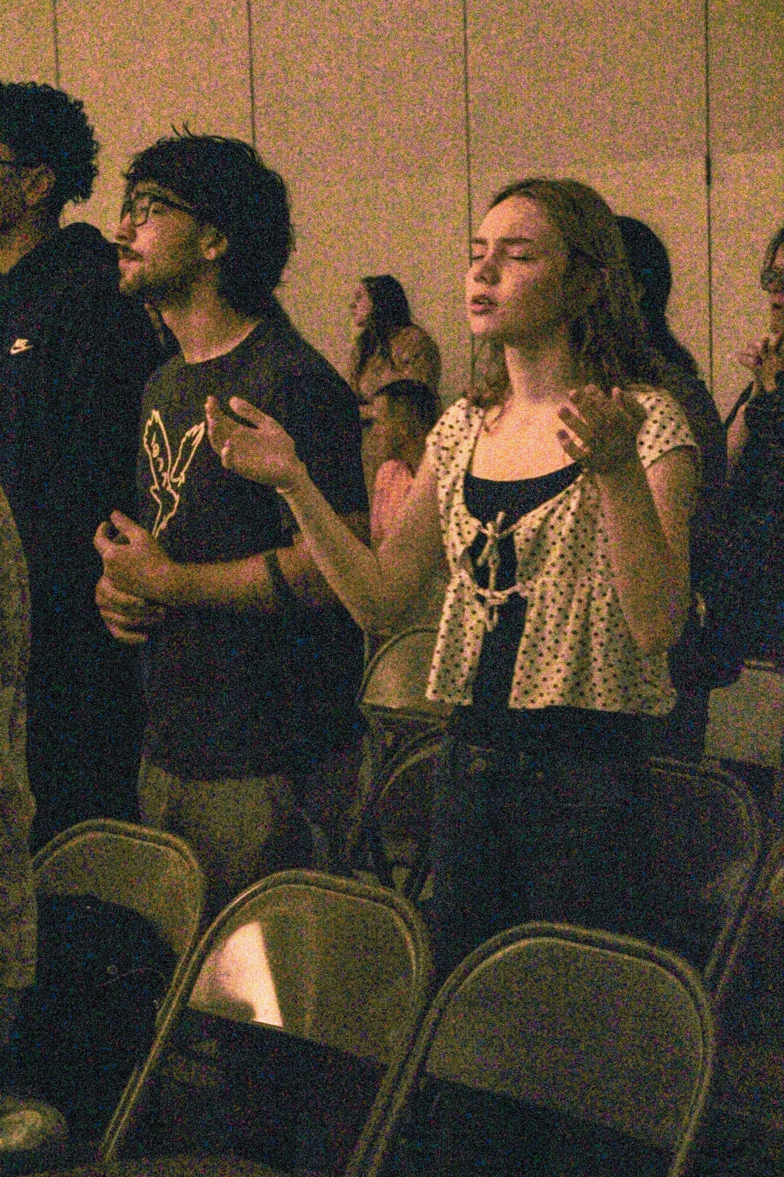 People standing in a dimly lit room, some are talking and one woman has her hand on her chest, with rows of empty seats in the foreground.