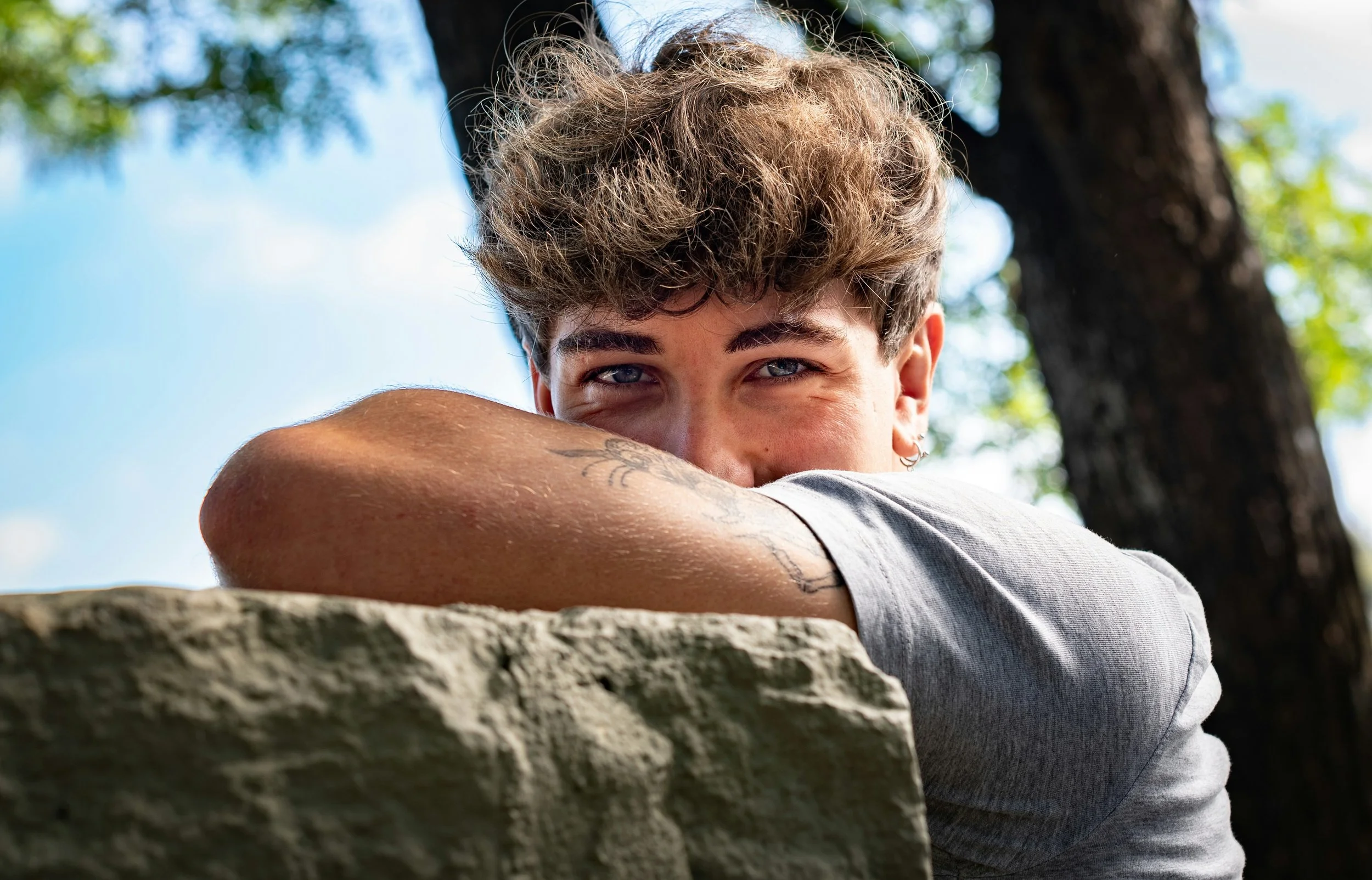A young person leaning on a stone wall outdoors, smiling and looking at the camera with a background of blue sky and green trees.