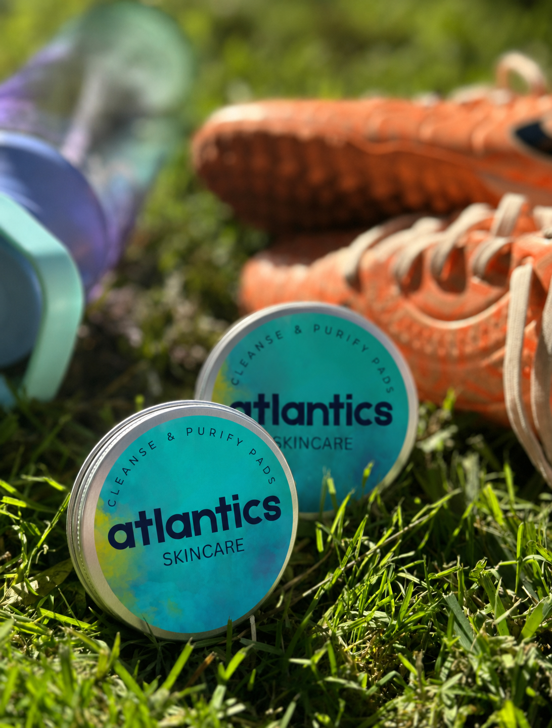 Two tropical-themed tins of Atlantic skincare products on grass with a pair of orange and white sneakers and a colorful water bottle in the background.