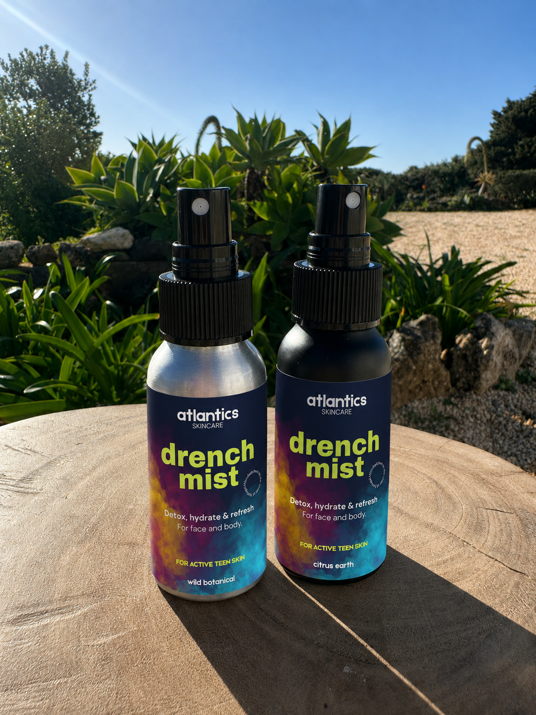 Two bottles of Atlantics skincare drench mist spray placed on a wooden surface outdoors with green plants and clear blue sky in the background.