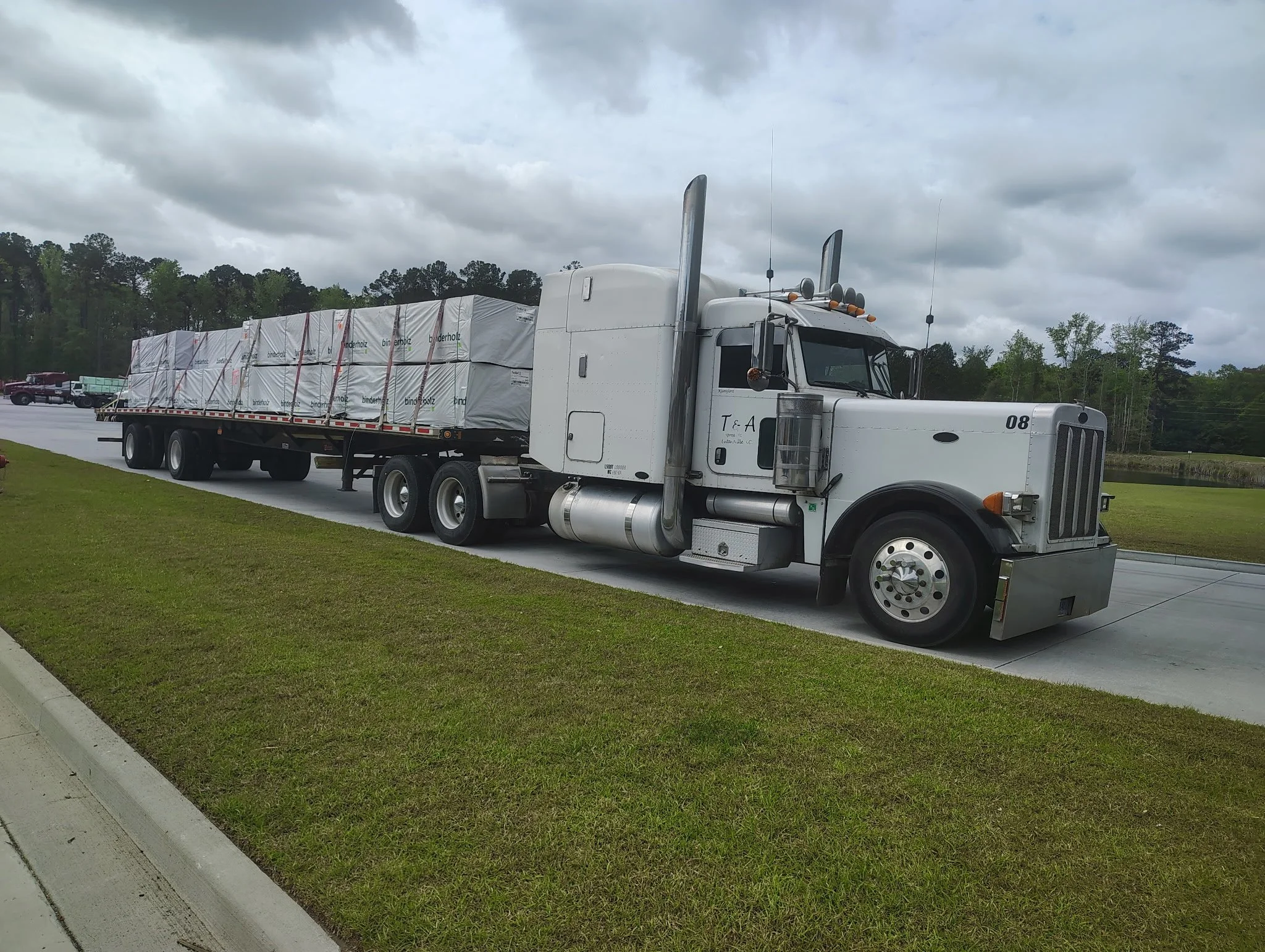 A white semi-truck hauling a flatbed trailer loaded with wrapped bricks, parked on the side of a road with cloudy sky and trees in the background.