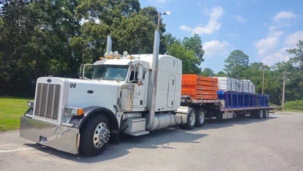 A semi-truck with a white cab and orange and blue cargo on a flatbed trailer parked on the side of a road, with trees and a blue sky in the background.
