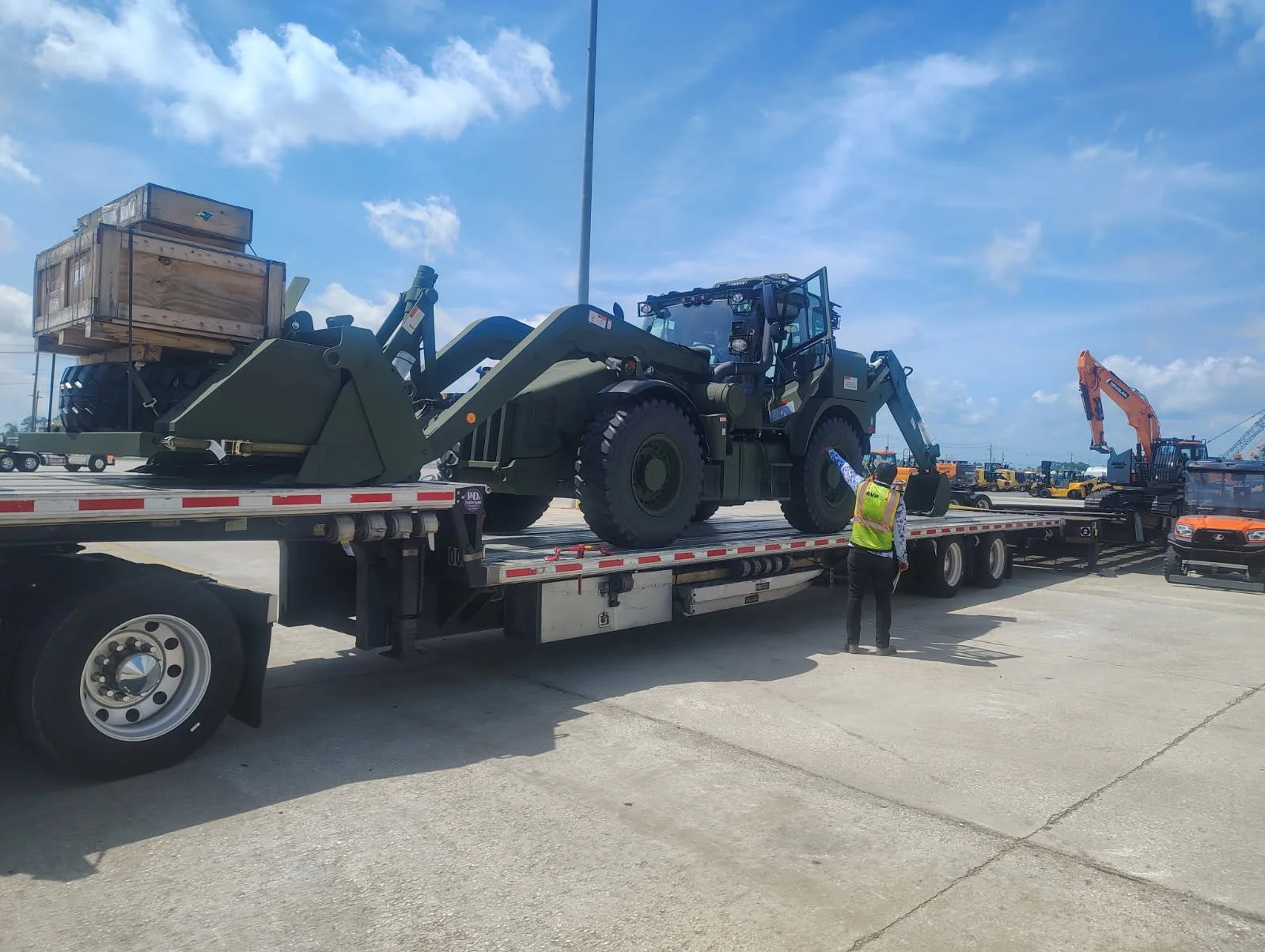 A large military green bulldozer with a wooden crate on top of its blade, loaded on a flatbed truck. A person wearing a yellow safety vest and a hard hat stands next to the truck, observing the equipment.
