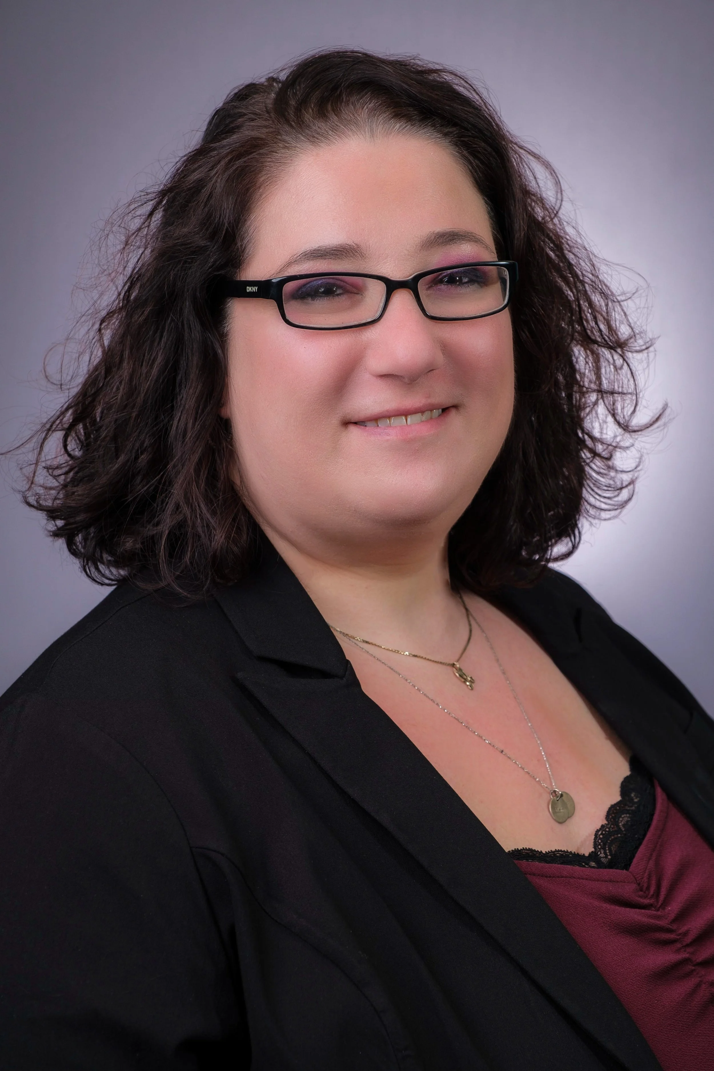 Carrie Rainen, a woman with curly dark hair, wearing glasses, a black blazer, and a burgundy top with lace detail, smiling at the camera.