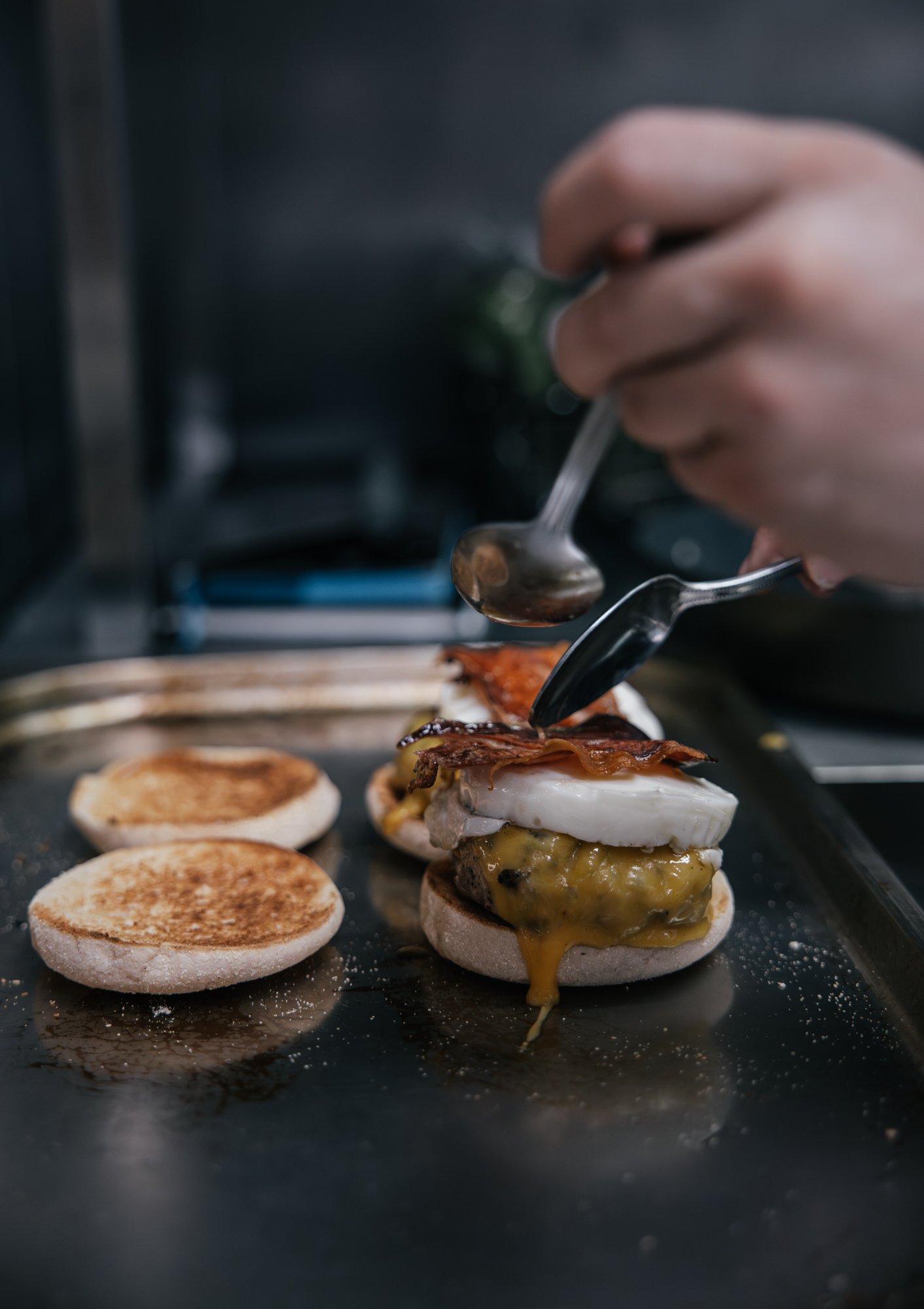 A close-up of a hand holding a spoon and adding sauce onto mini burgers on a baking tray, with some toasted buns in the background.