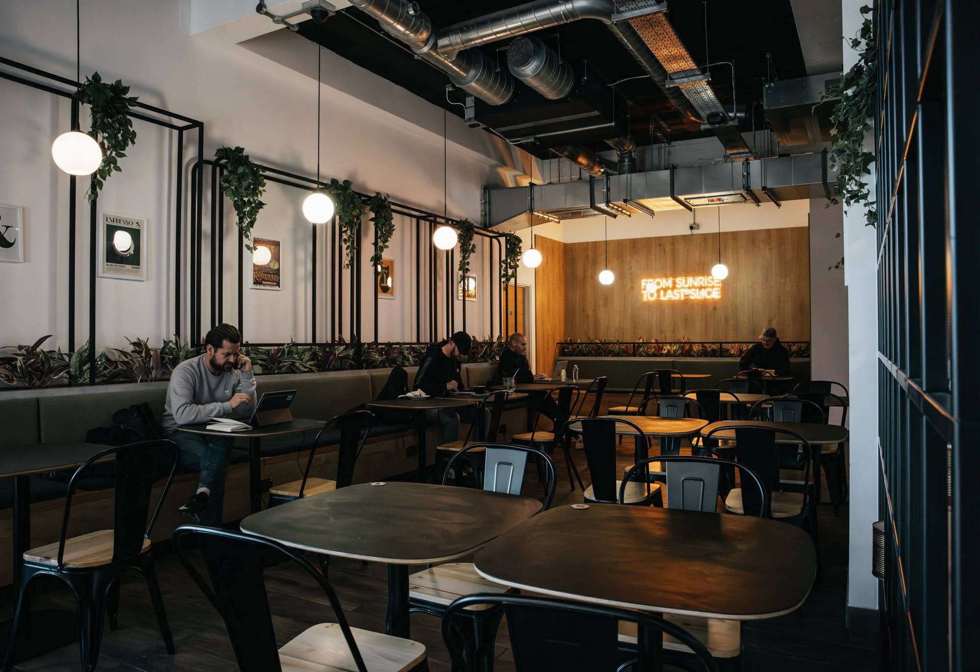 Interior of a modern coffee shop with hanging globe lights, framed art on white walls, plants decorating the space, and a neon sign on a wooden wall saying "From sunrise to last slice." Several people are seated at tables, working or using devices.