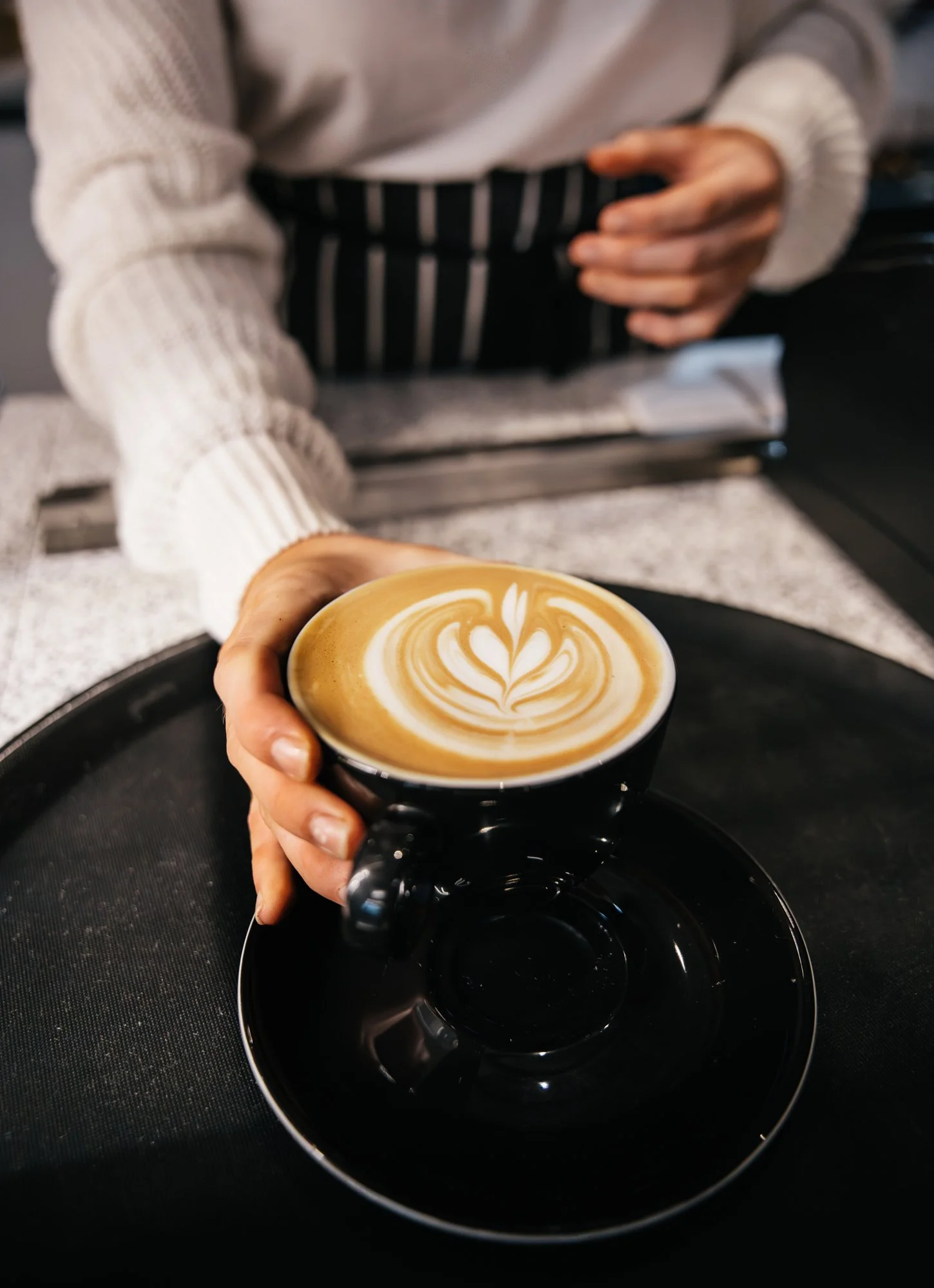 A person holding a black coffee cup with latte art, sitting at a counter with a black tray underneath the cup.