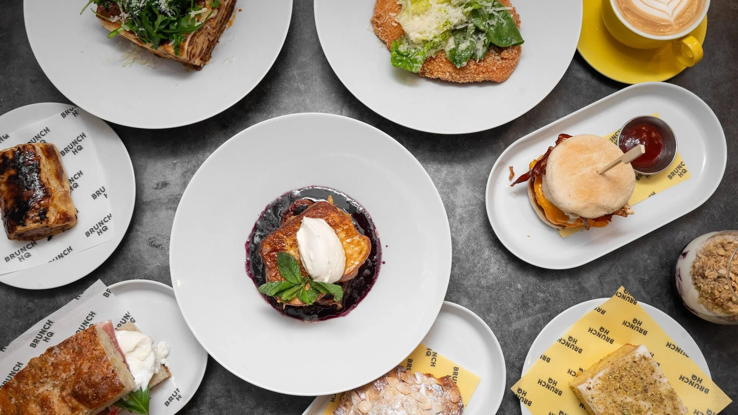 Variety of desserts and small food items on white plates, including a slice of cake, a fruit tart, a ham sandwich, a chicken sandwich, and a pastry, arranged on a dark surface.