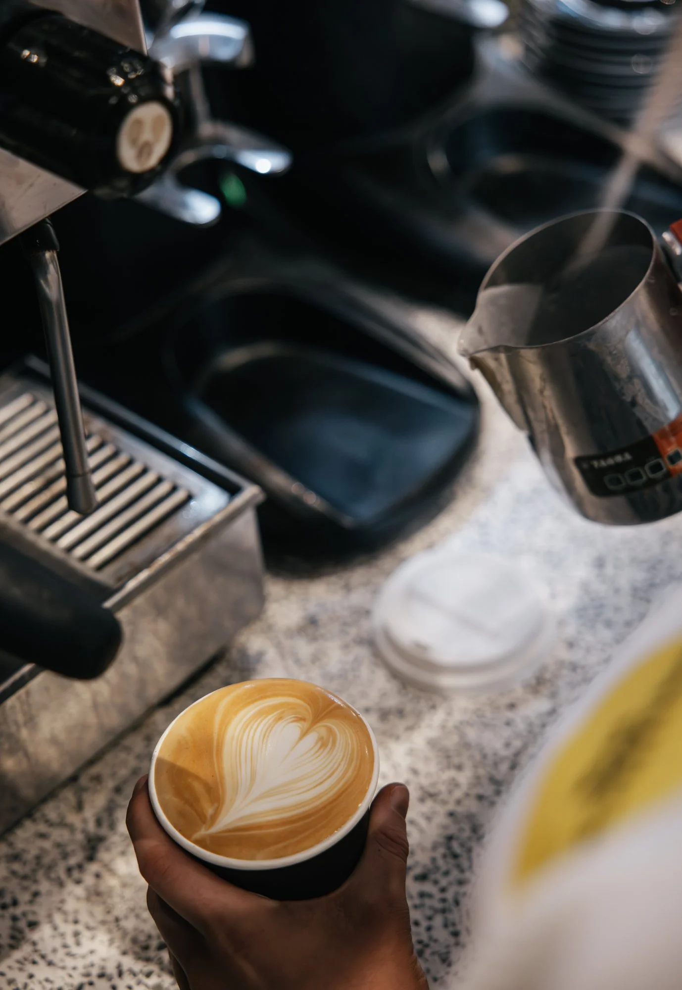 Person holding a cup of coffee with latte art, in front of a coffee machine and coffee cups.