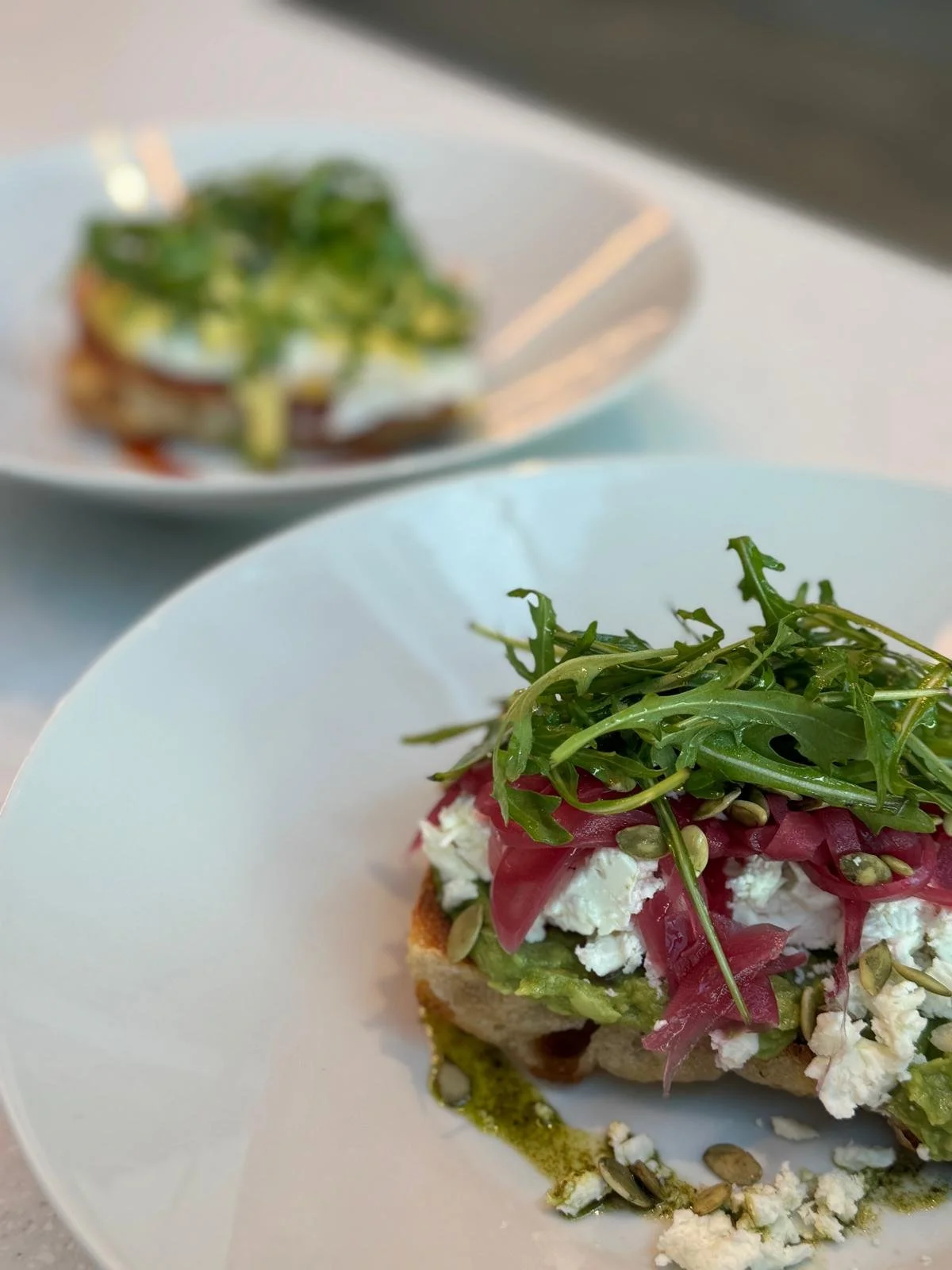 Close-up of an open-faced sandwich with arugula, ricotta cheese, sun-dried tomatoes, and pumpkin seeds on bread, with a blurred salad in the background.